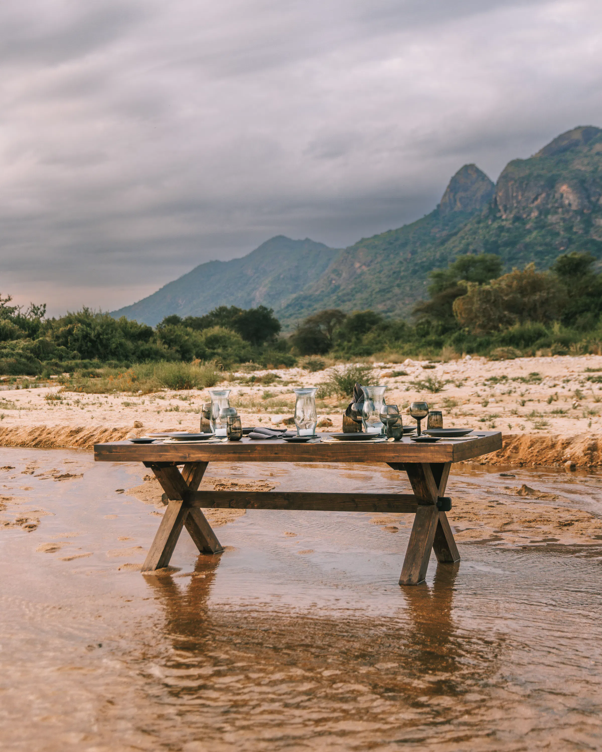 A dining table is set in a shallow riverbed at Kalepo, with mountains and cloudy skies stretching beyond.