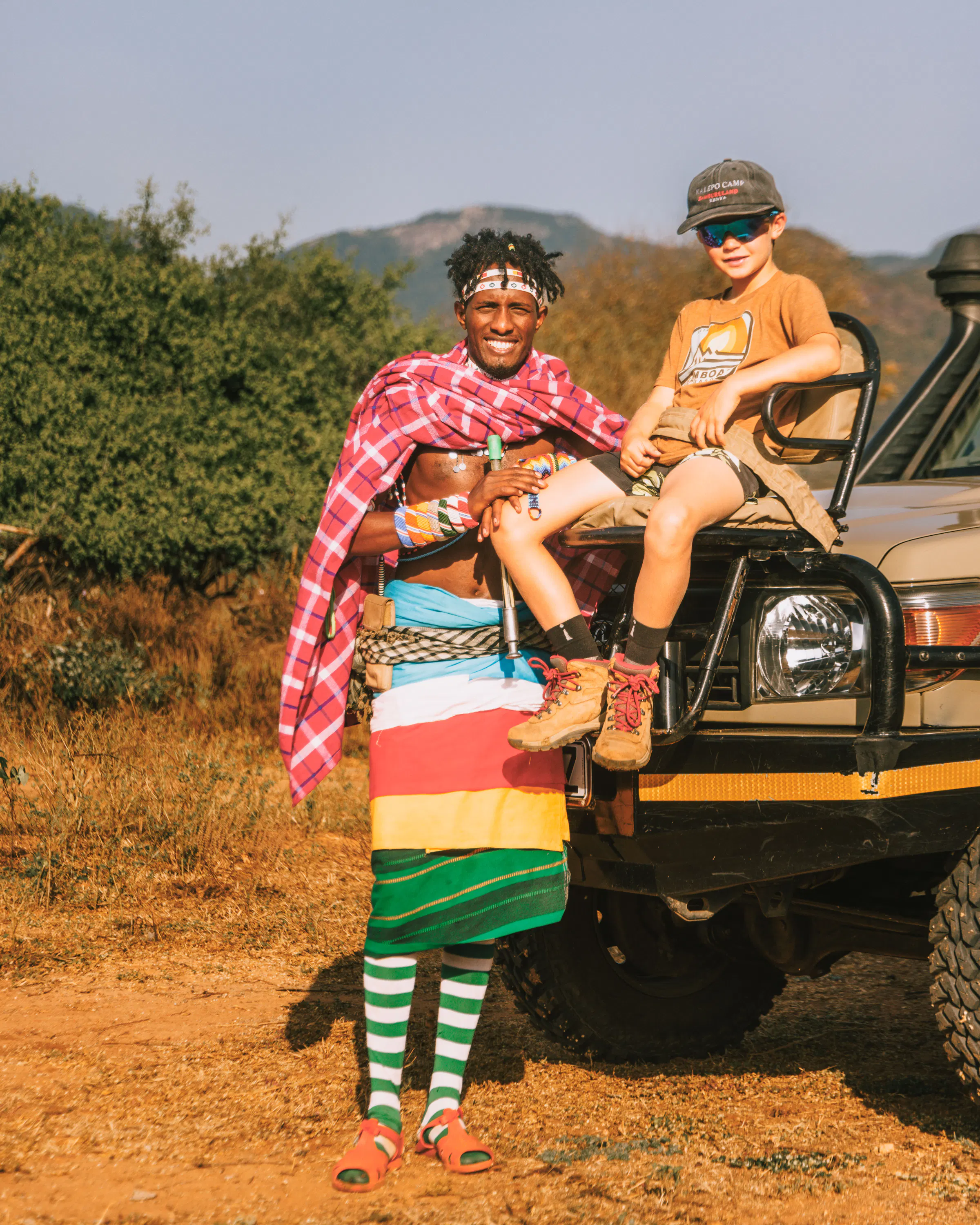 A Samburu guide stands beside a child on a safari vehicle at Kalepo, with dry hills and shrubs around them.