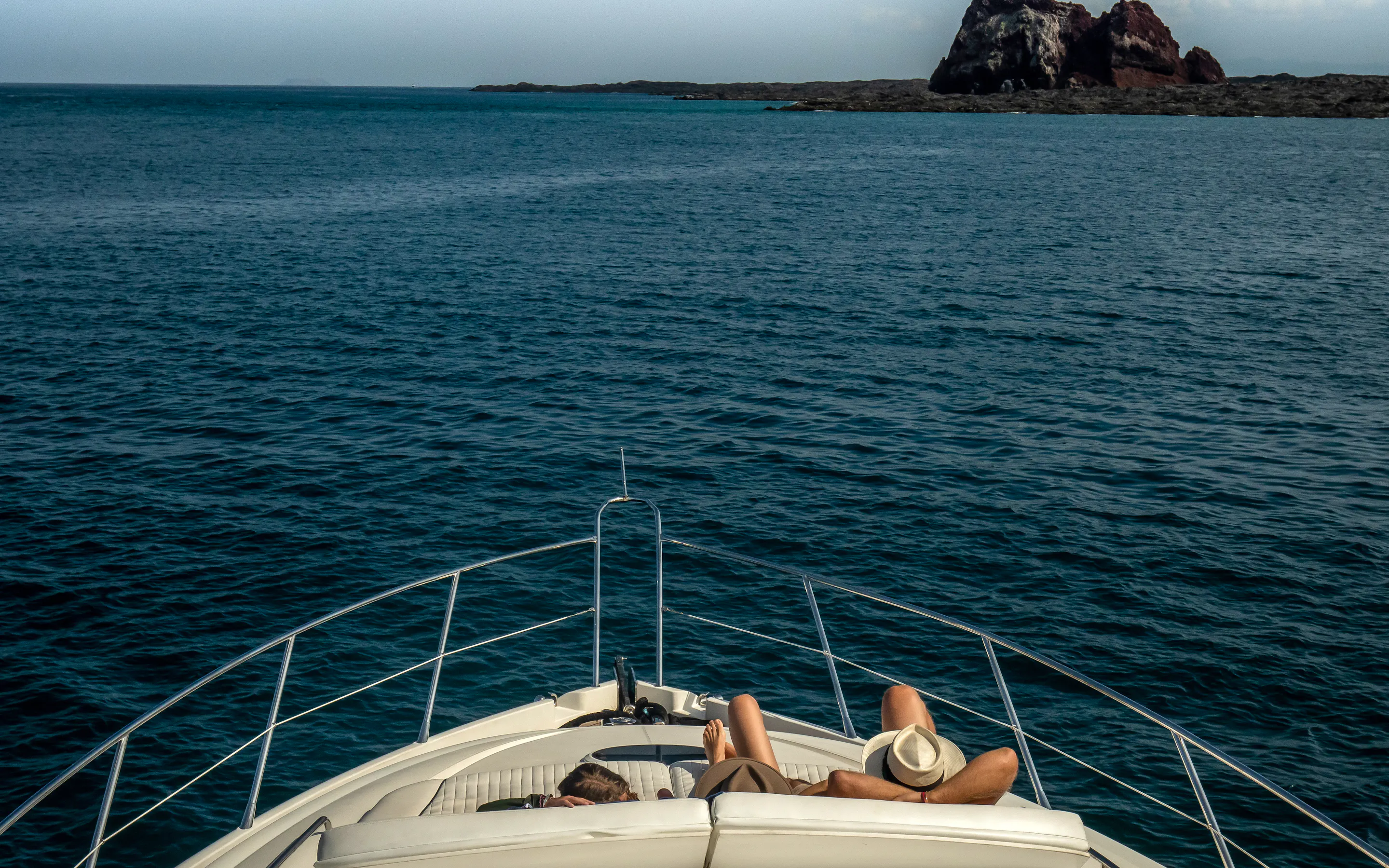 Two guests recline on the bow of a yacht approaching rocky islets off Santa Cruz in the Galapagos Islands.