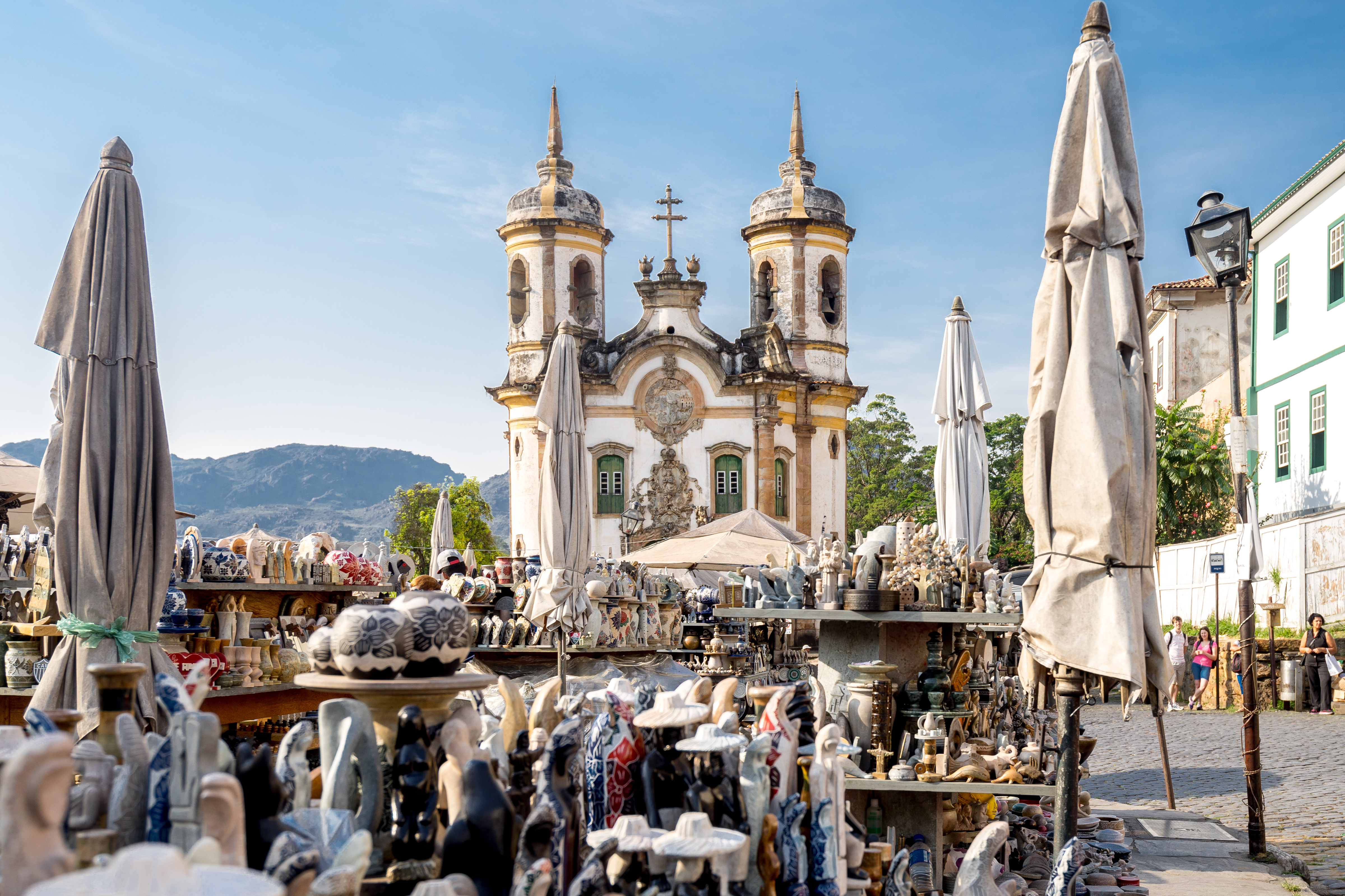 A church rises beyond an artisan market in Ouro Preto, with ceramic stalls and mountains under a clear sky.