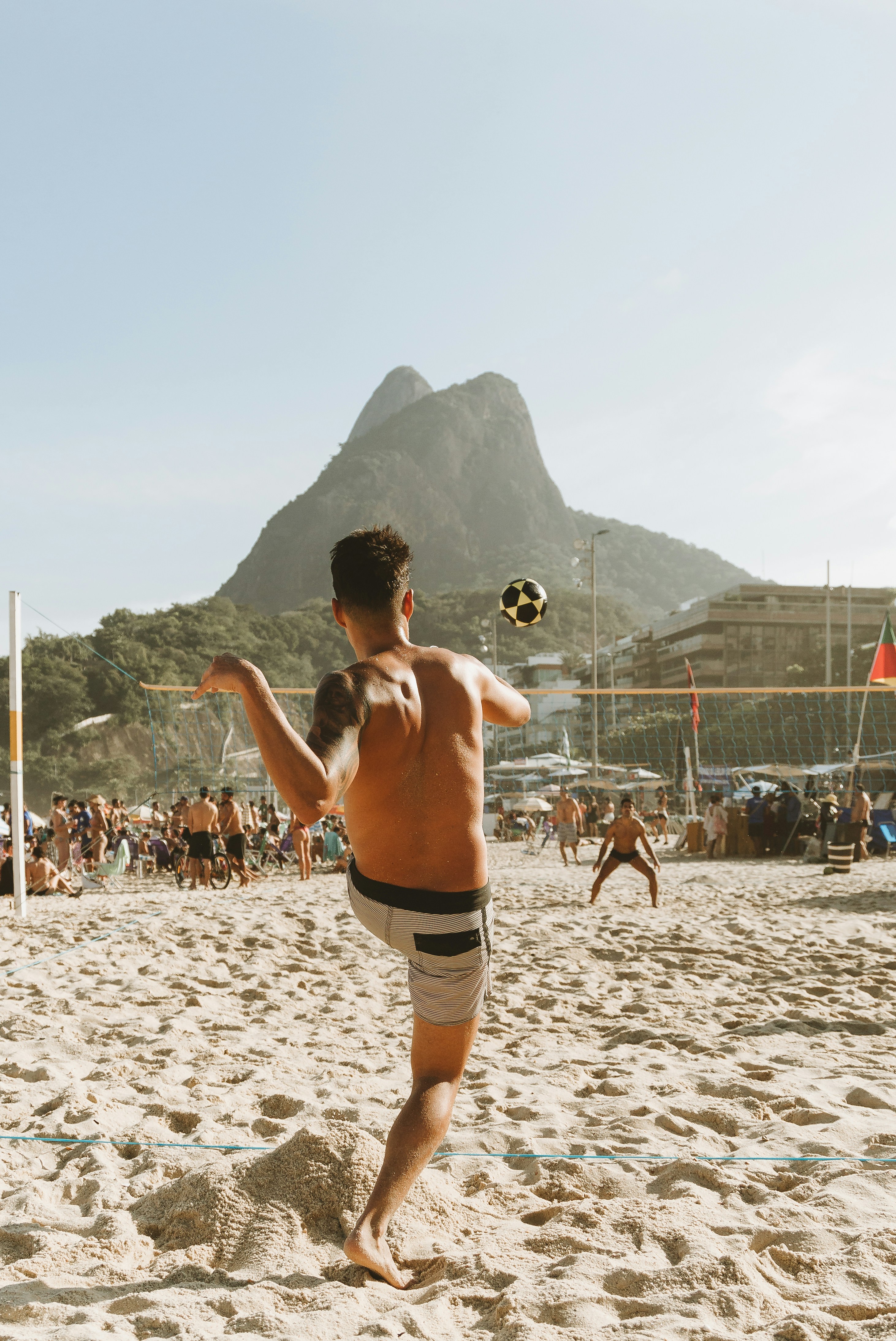 A beach player tosses a ball on Rio's sand, with Dois Irmãos rising in the background under bright afternoon light.