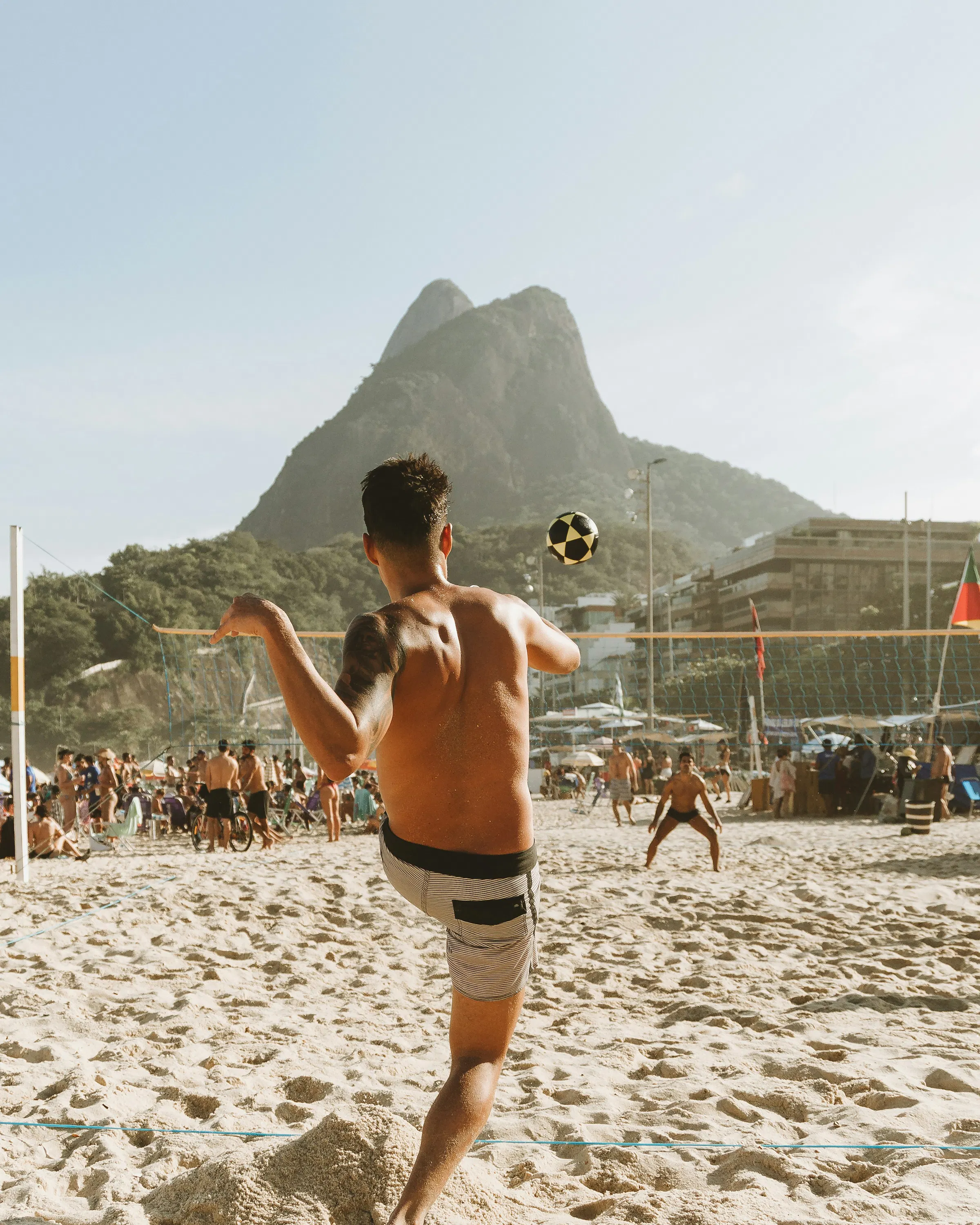 A beach player tosses a ball on Rio's sand, with Dois Irmãos rising in the background under bright afternoon light.