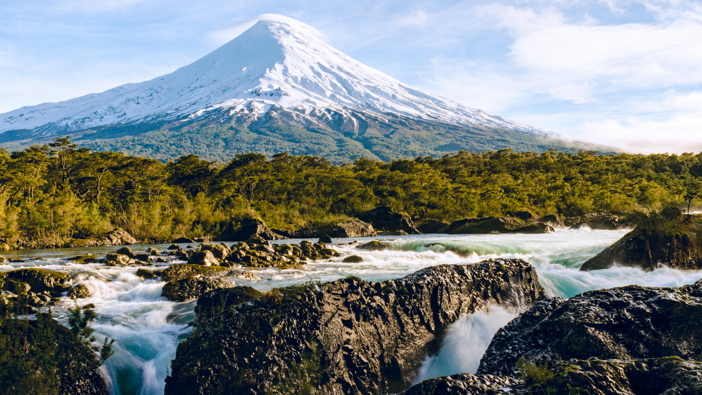 Snow-capped Osorno Volcano rises above rushing black-rock waterfalls and dense forest beneath a bright blue sky.