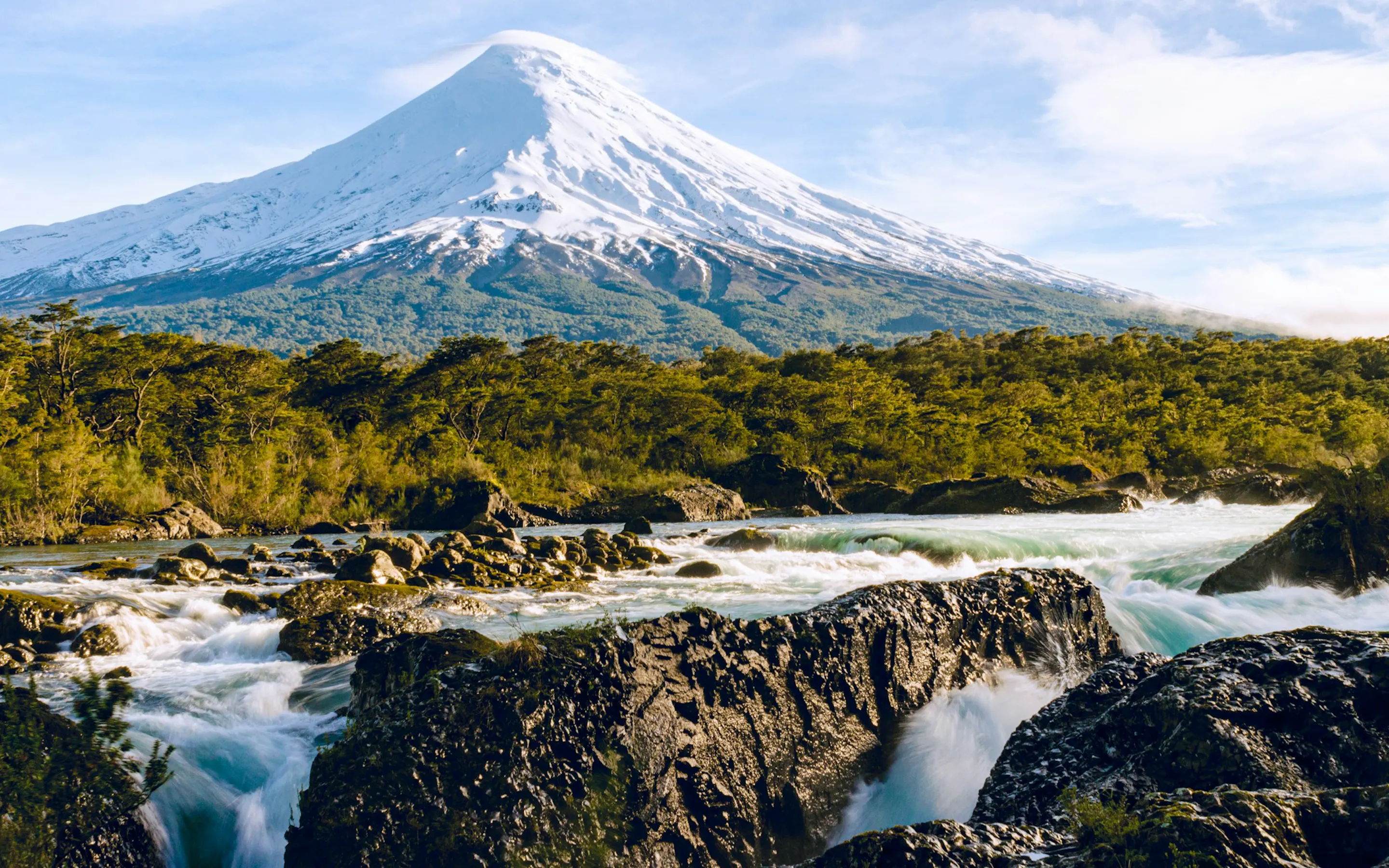 Snow-capped Osorno Volcano rises above rushing black-rock waterfalls and dense forest beneath a bright blue sky.