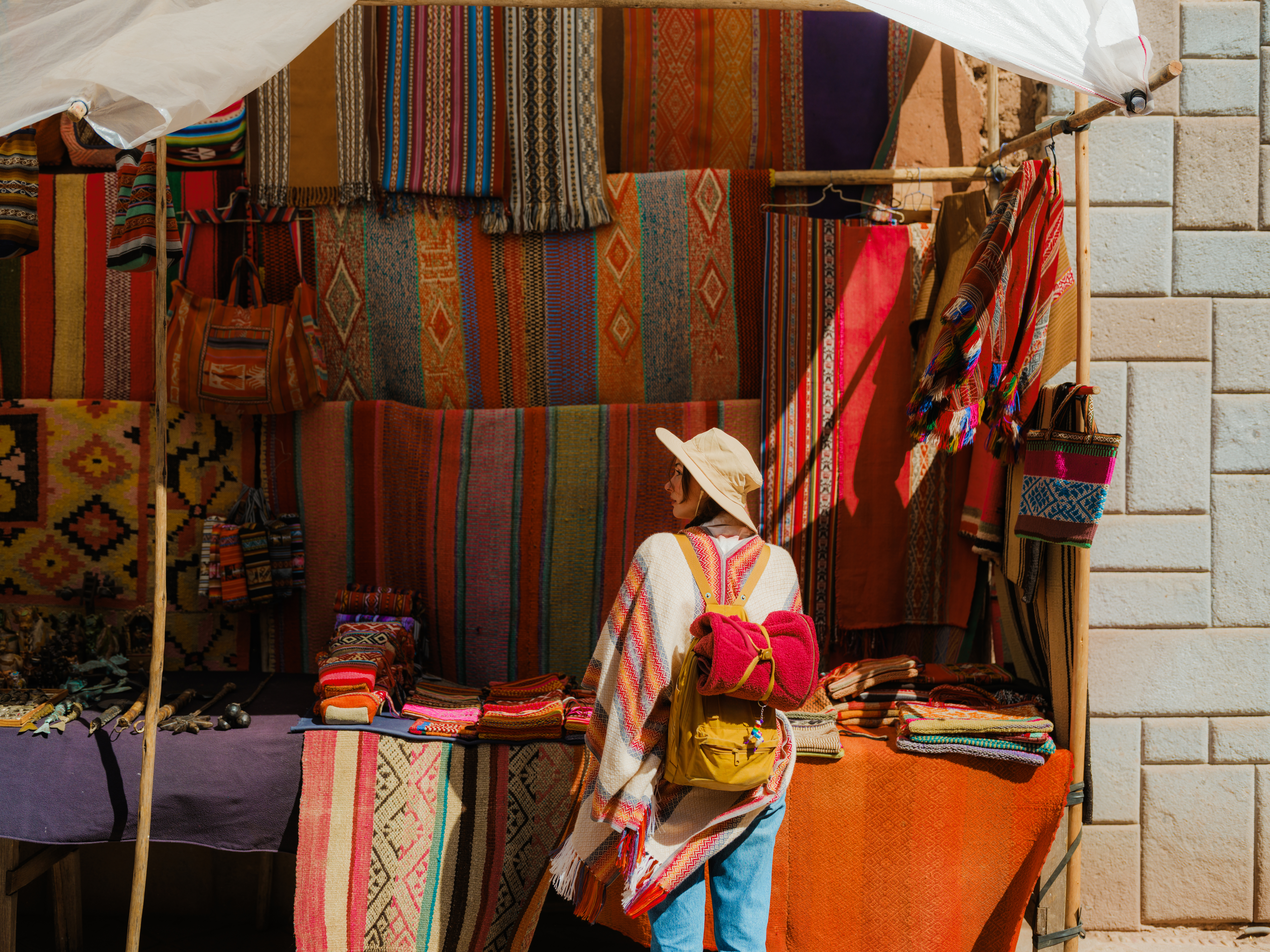 A woman in a white hat browses woven textiles and souvenirs at a colorful market stall in Pisac, Peru's Sacred Valley.