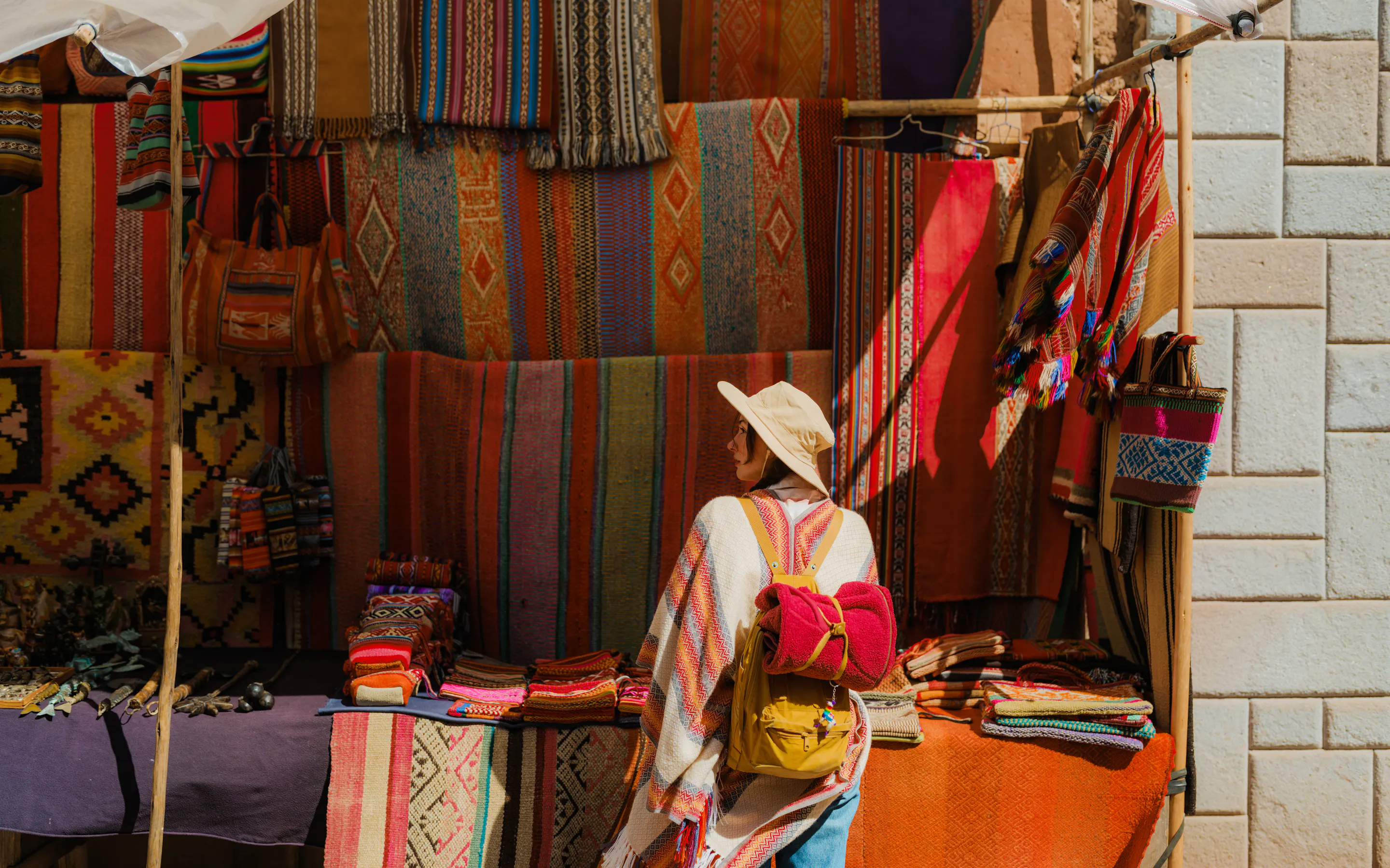 A woman in a white hat browses woven textiles and souvenirs at a colorful market stall in Pisac, Peru's Sacred Valley.