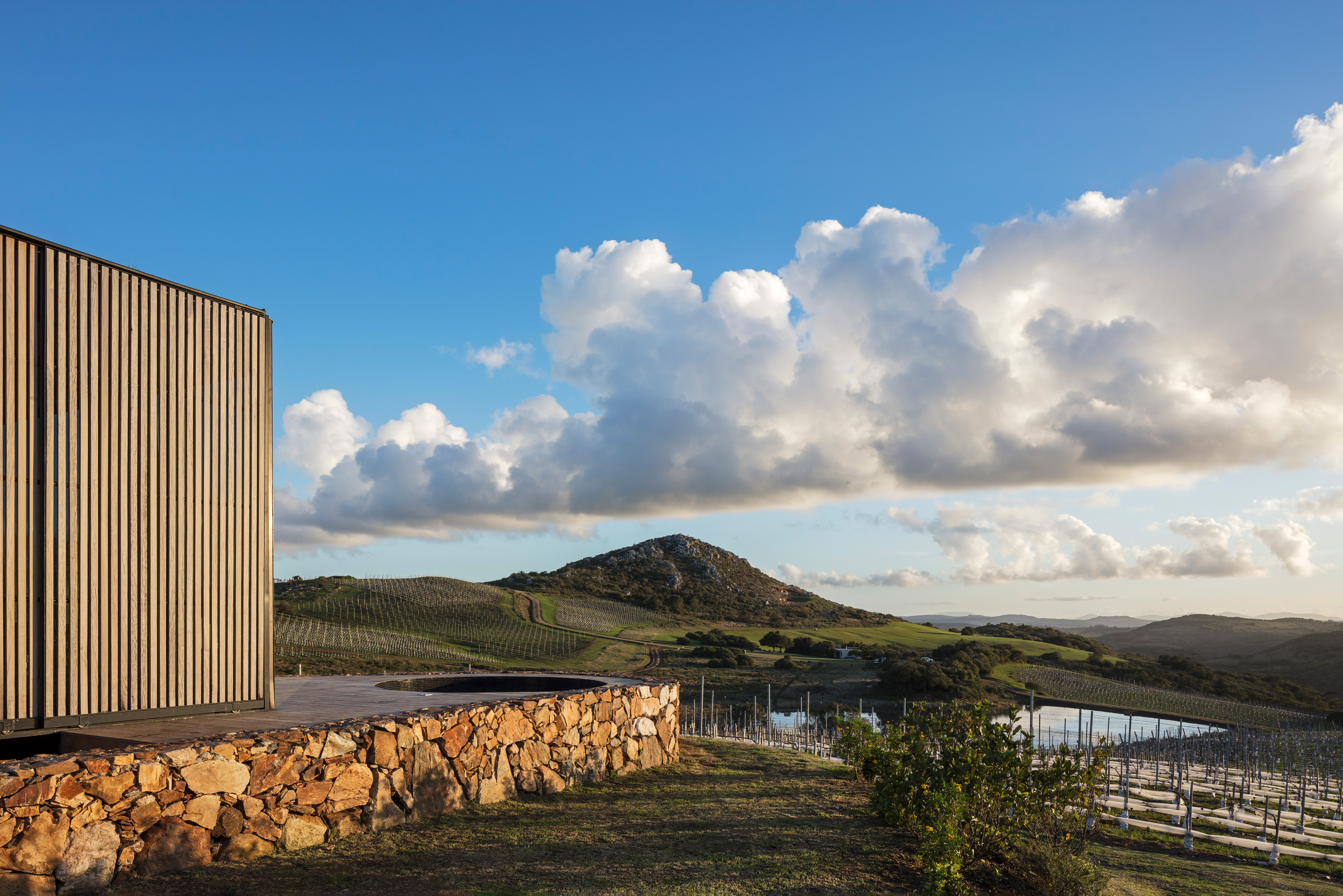 A modern hillside cabin with stone walls overlooks rolling vineyards and dramatic clouds at Sacromonte in Uruguay.