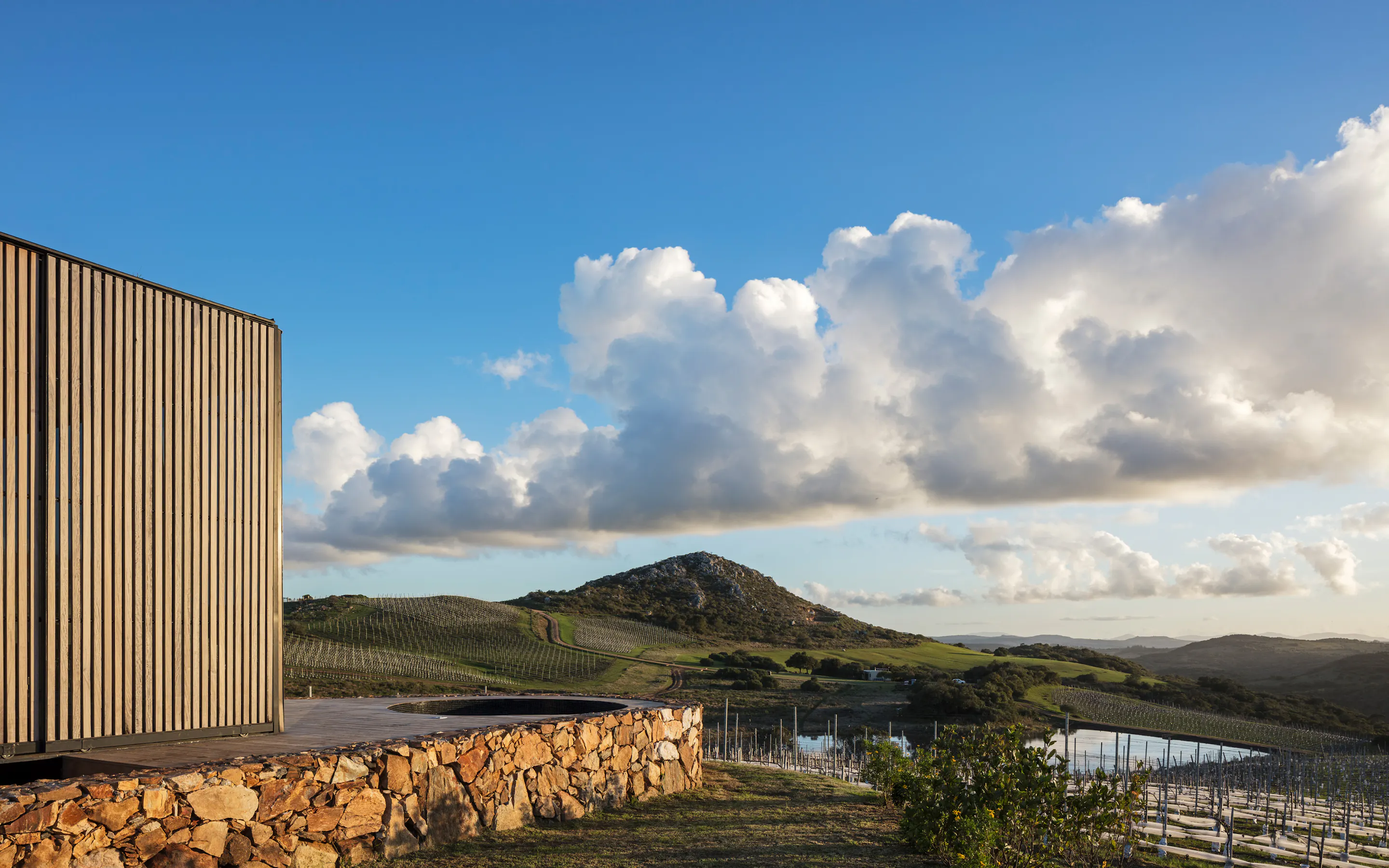A modern hillside cabin with stone walls overlooks rolling vineyards and dramatic clouds at Sacromonte in Uruguay.