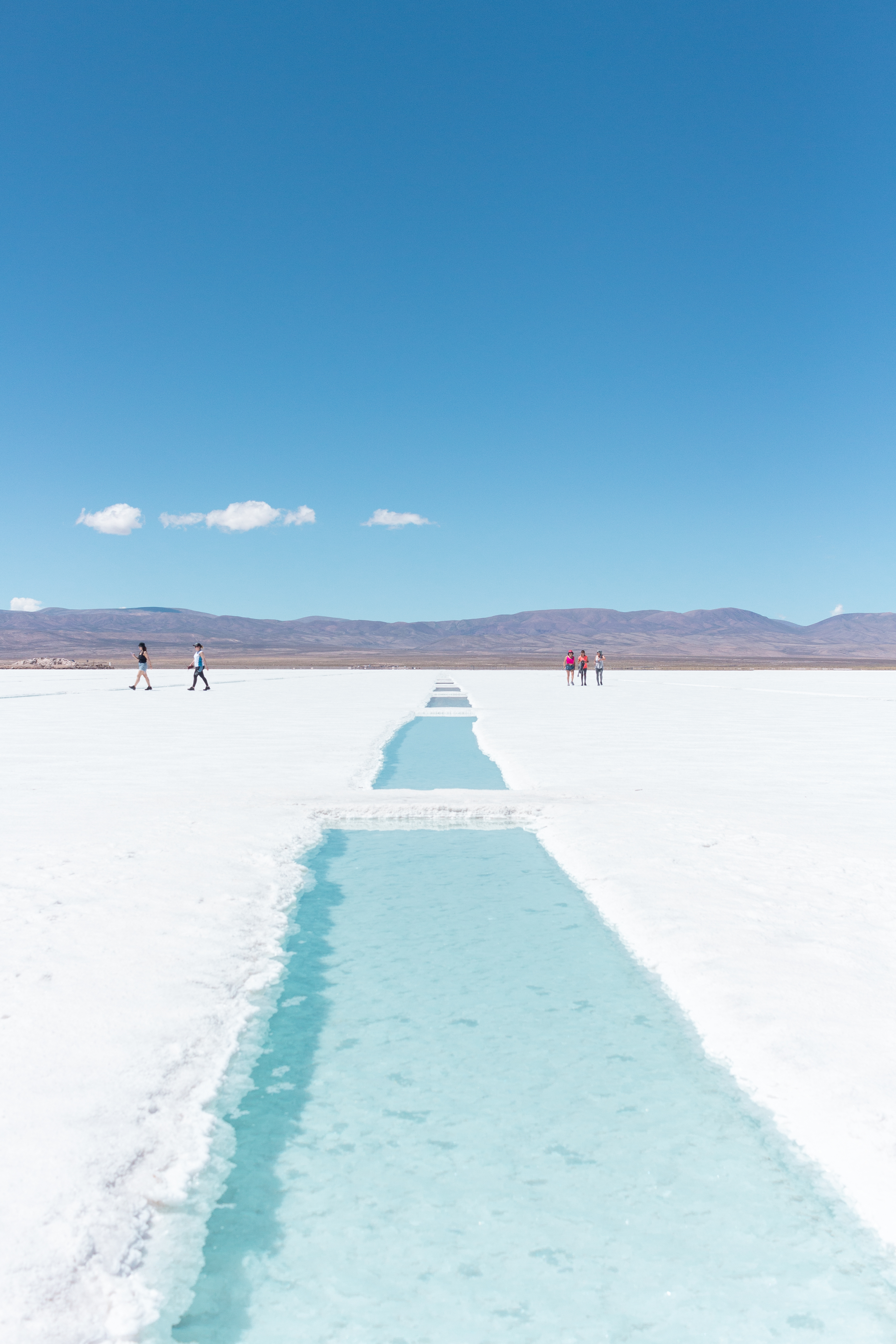 A turquoise salt pool cuts through the white flats of Salinas Grandes as visitors cross beneath a blue sky.