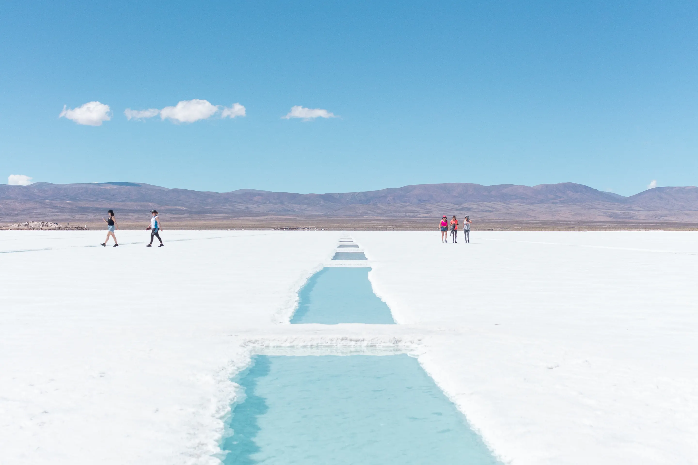 A turquoise salt pool cuts through the white flats of Salinas Grandes as visitors cross beneath a blue sky.