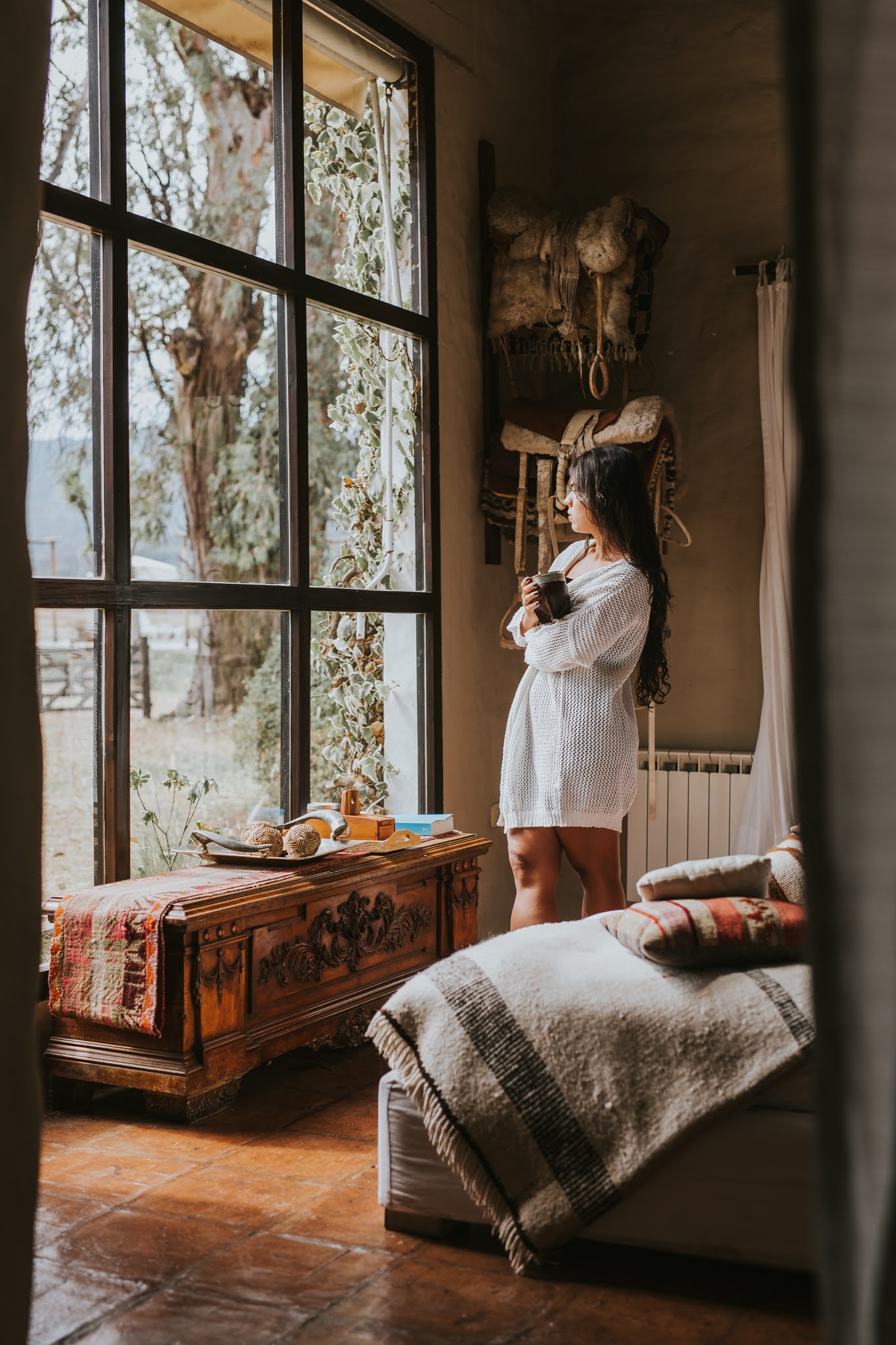 A guest stands by a tall window holding a cup in a rustic suite at House of Jasmines in northern Argentina.