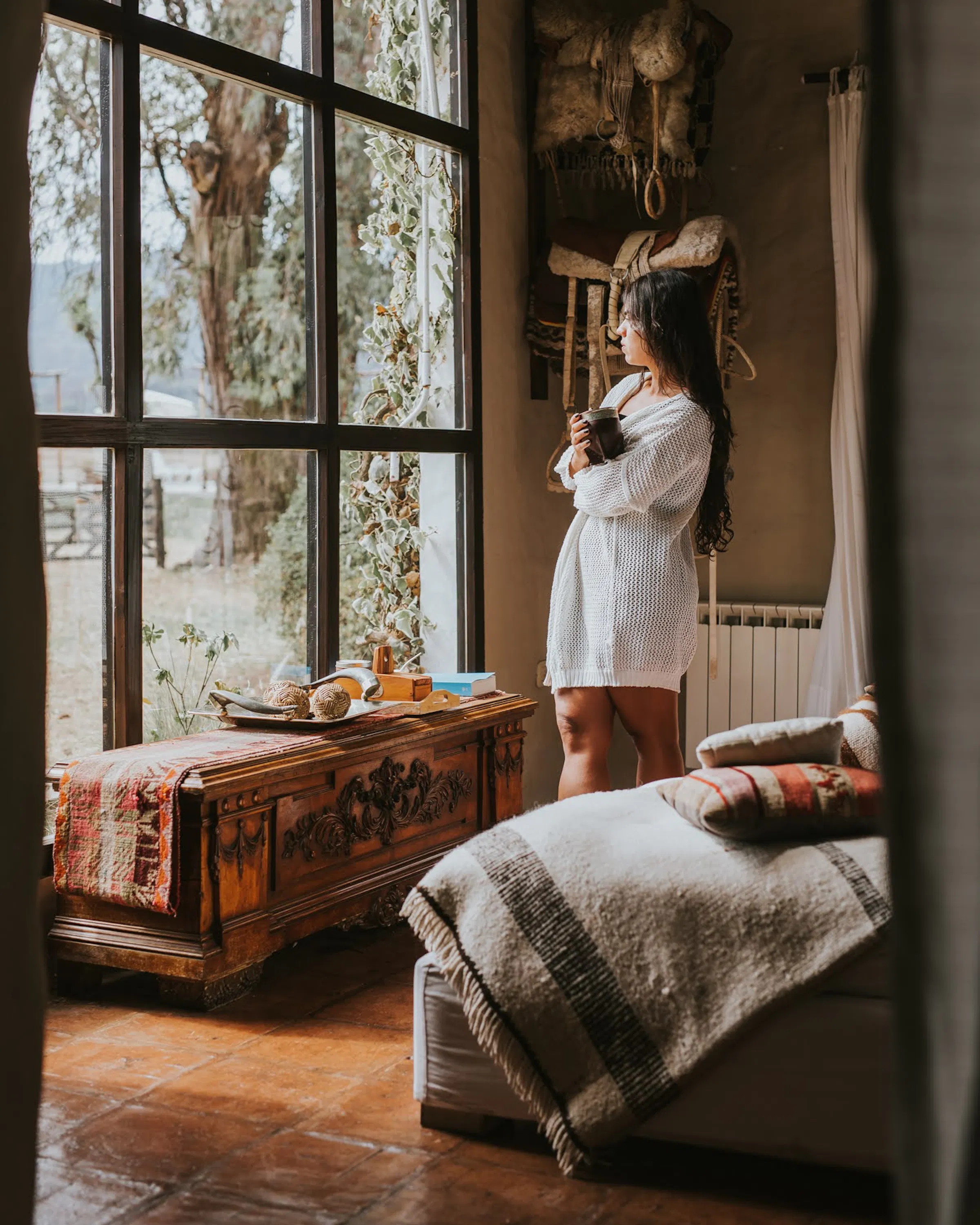 A guest stands by a tall window holding a cup in a rustic suite at House of Jasmines in northern Argentina.