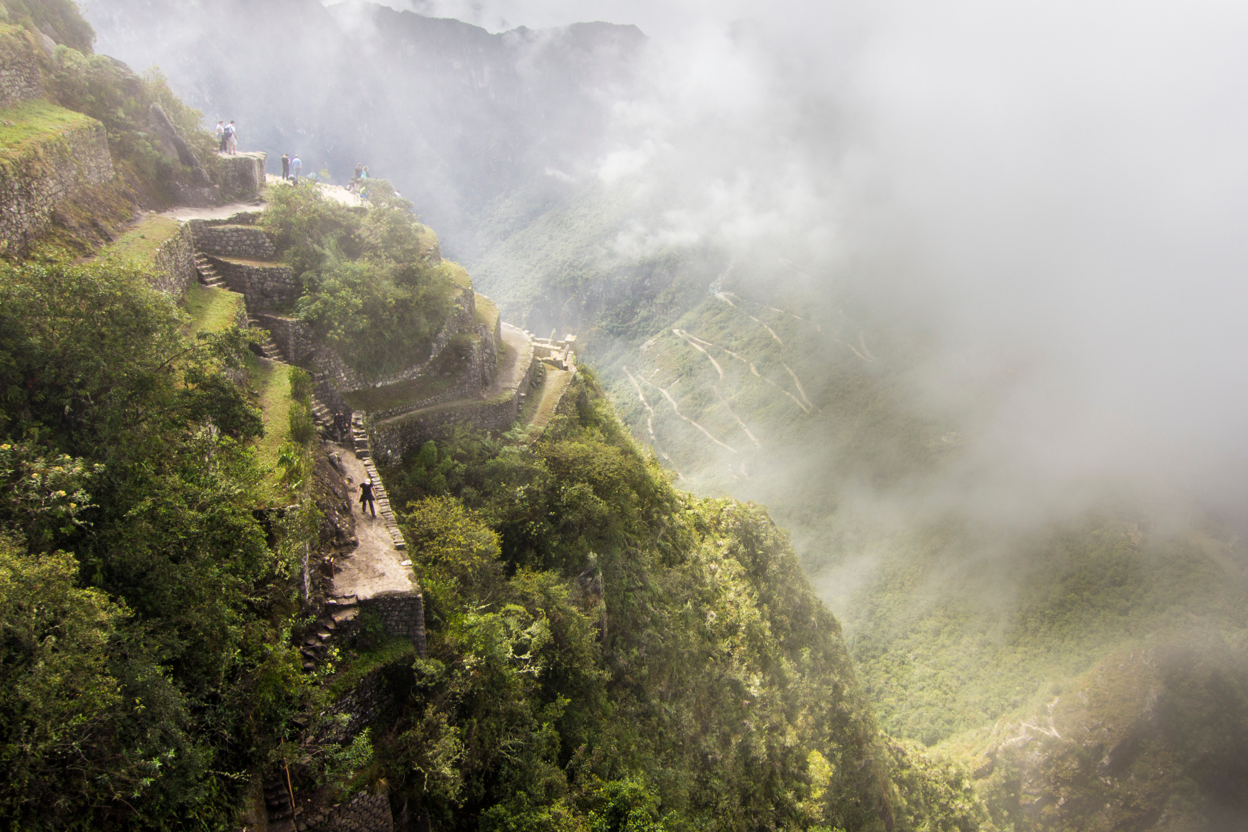 Misty terraces cling to the steep green slopes of Machu Picchu as visitors pause along the stone paths in soft light.
