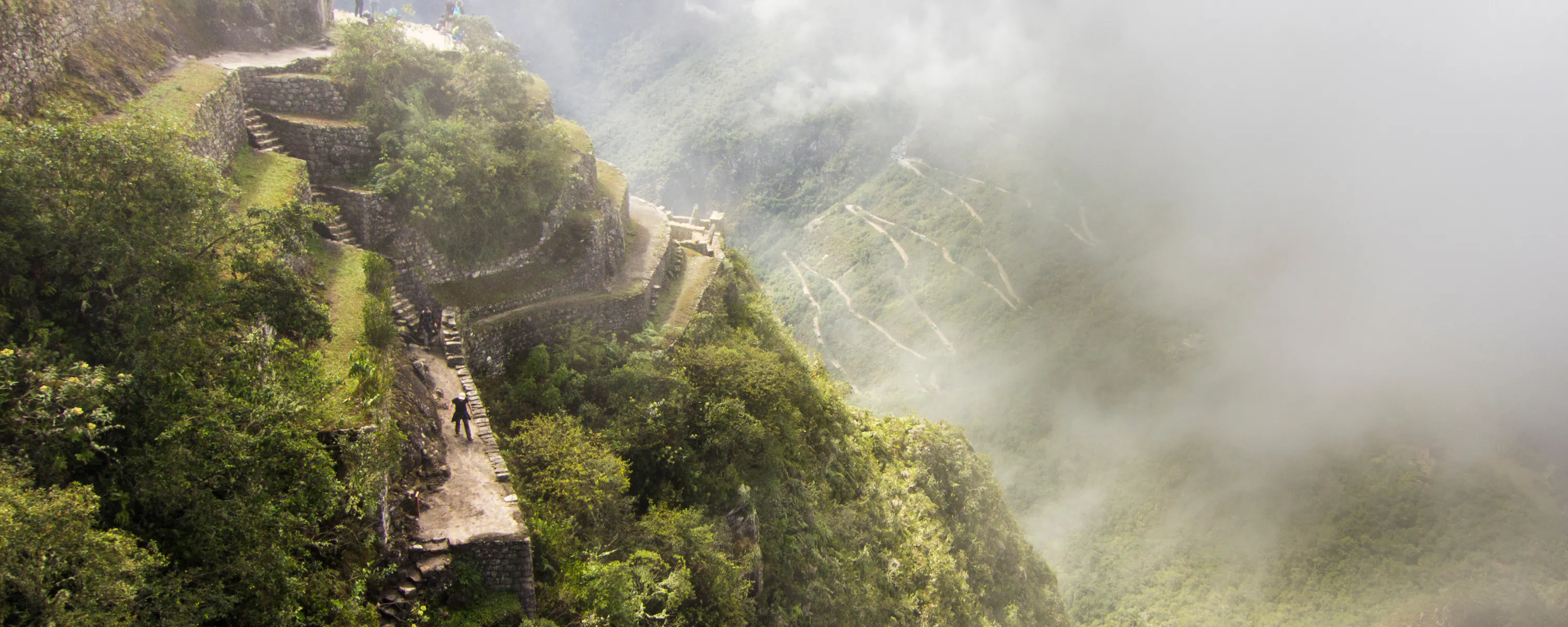 Misty terraces cling to the steep green slopes of Machu Picchu as visitors pause along the stone paths in soft light.