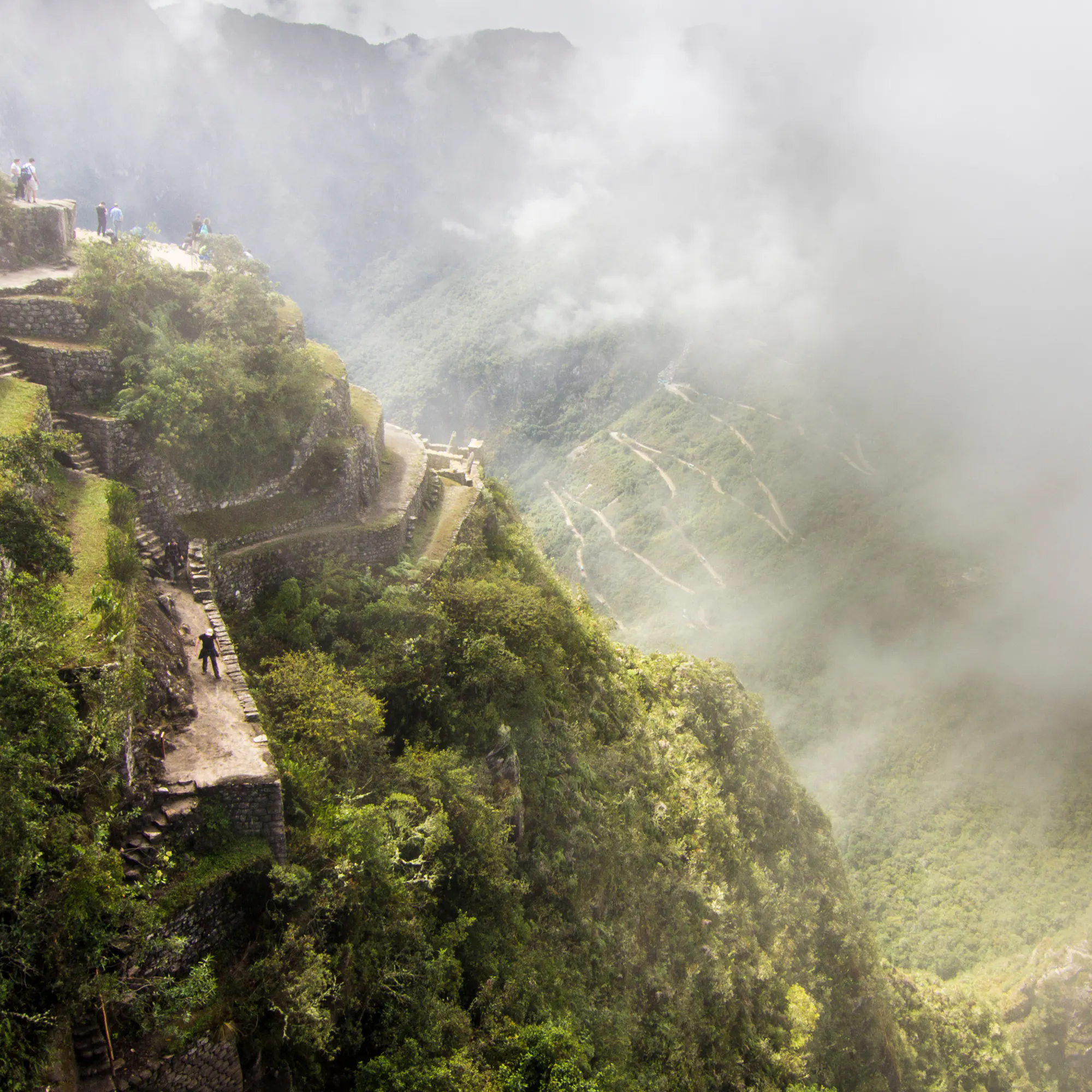 Misty terraces cling to the steep green slopes of Machu Picchu as visitors pause along the stone paths in soft light.