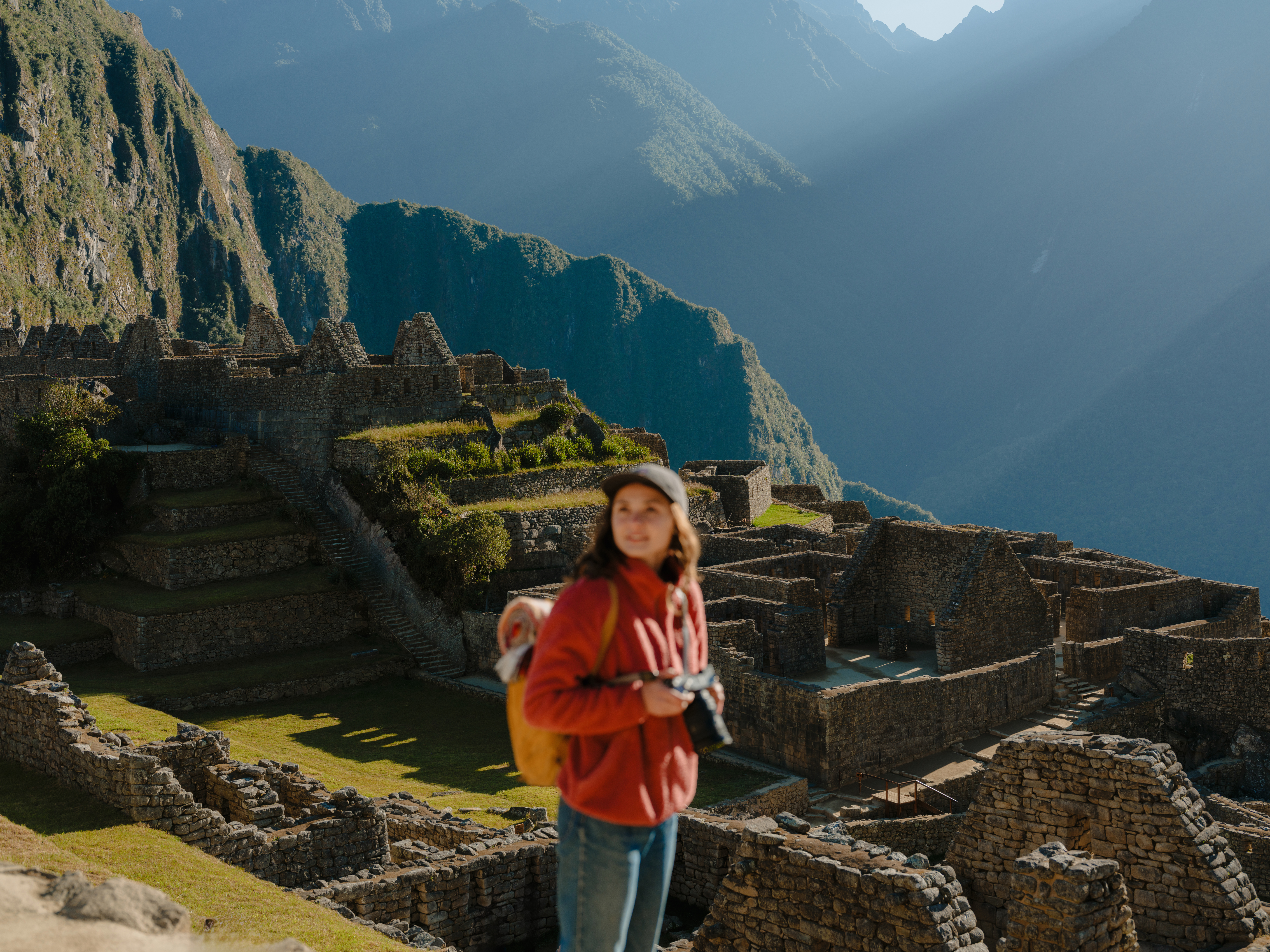 A traveler with a camera stands before the stone ruins of Machu Picchu beneath the steep Andean slopes at sunrise.