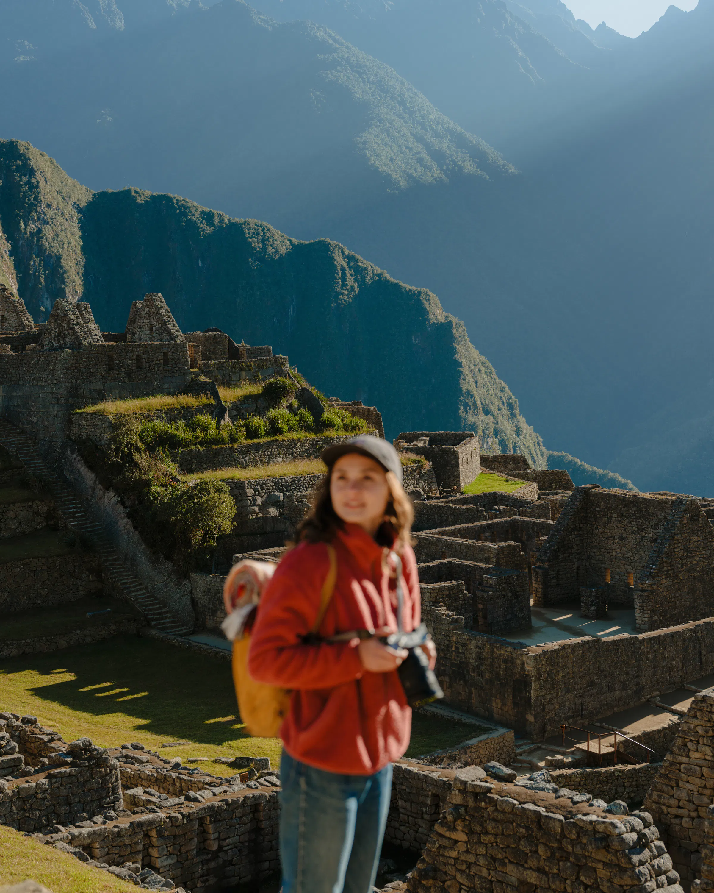 A traveler with a camera stands before the stone ruins of Machu Picchu beneath the steep Andean slopes at sunrise.