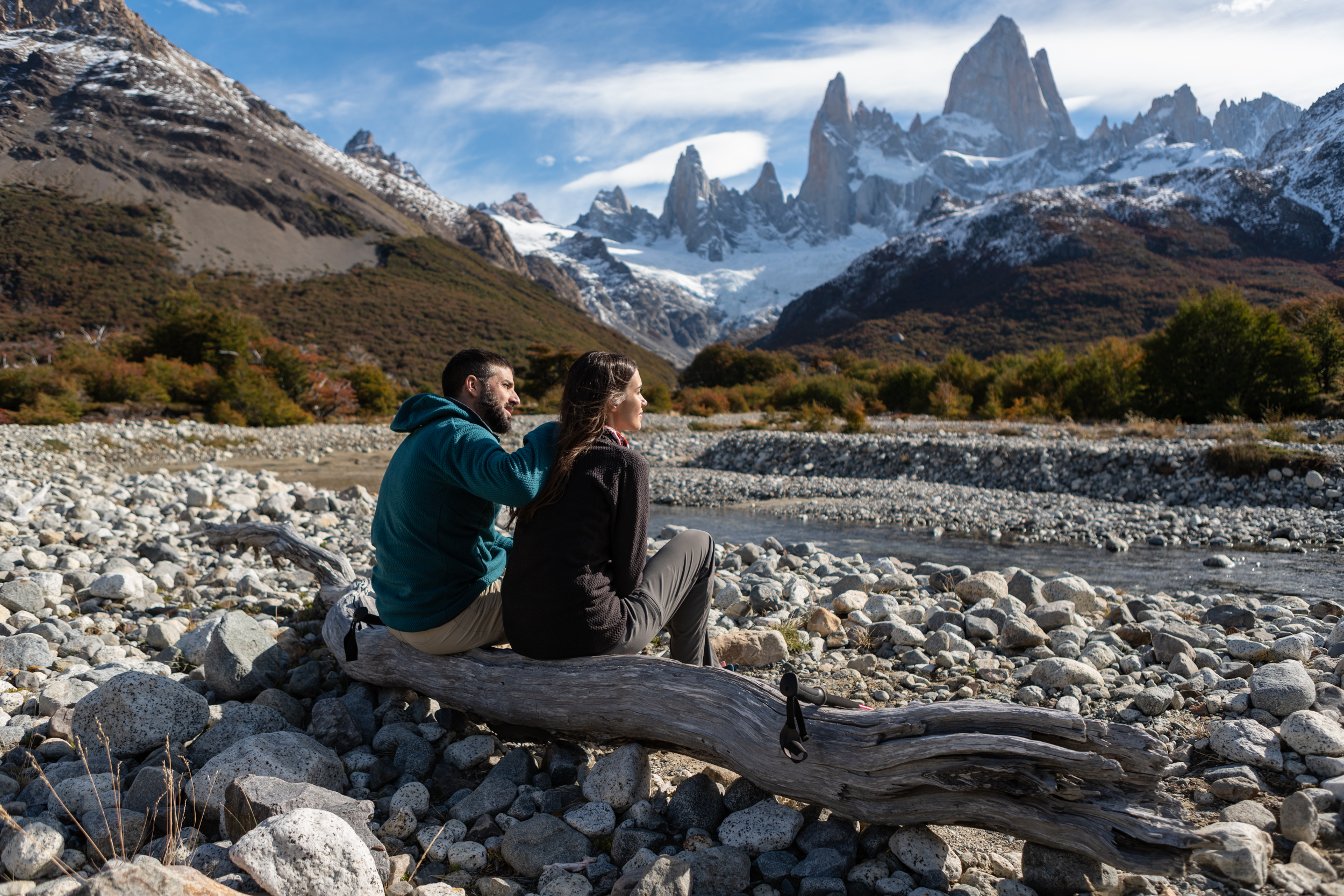 Two travelers sit by a rocky riverbank facing the snowy peaks and sharp spires near El Chalten, Argentina.