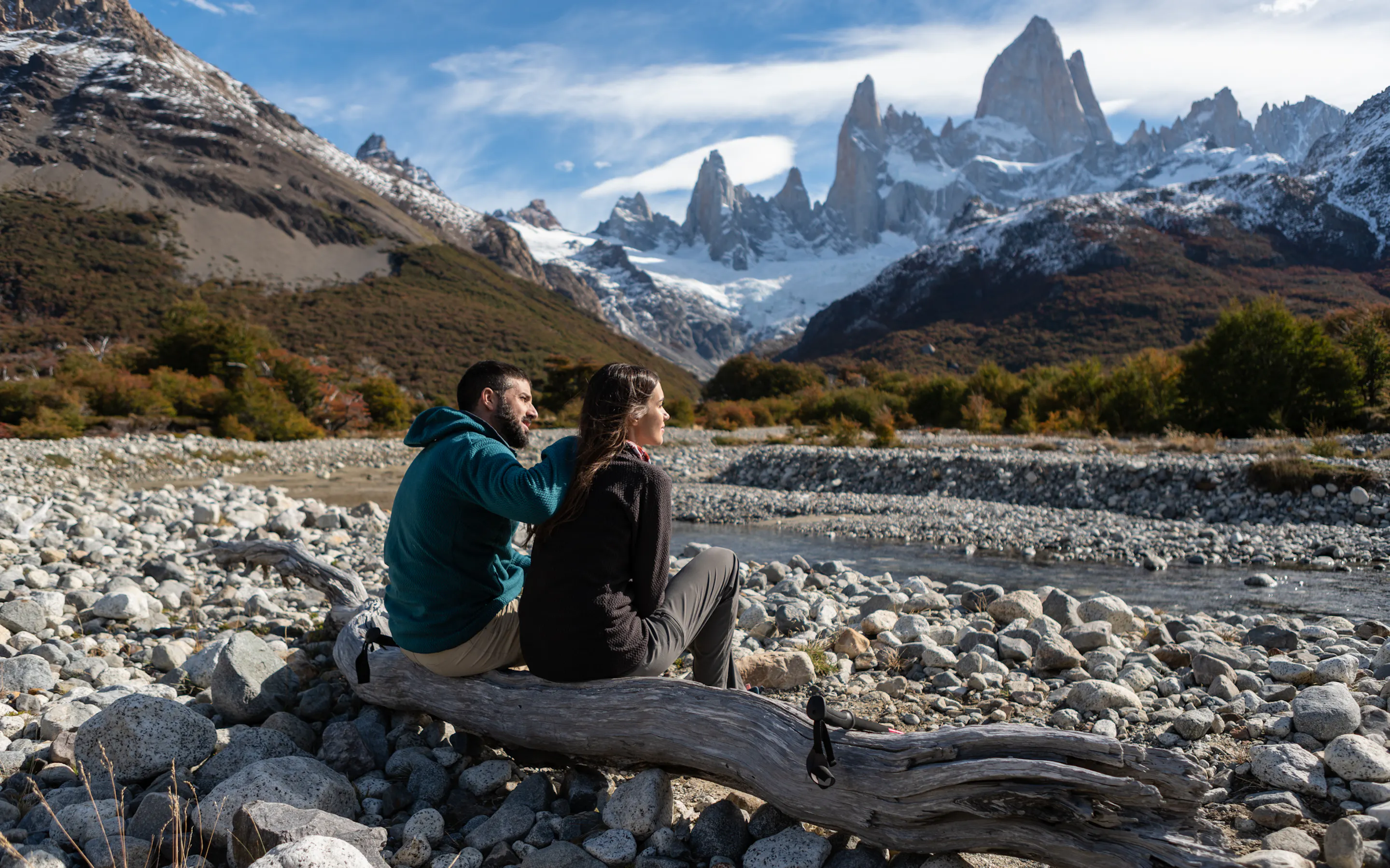 Two travelers sit by a rocky riverbank facing the snowy peaks and sharp spires near El Chalten, Argentina.