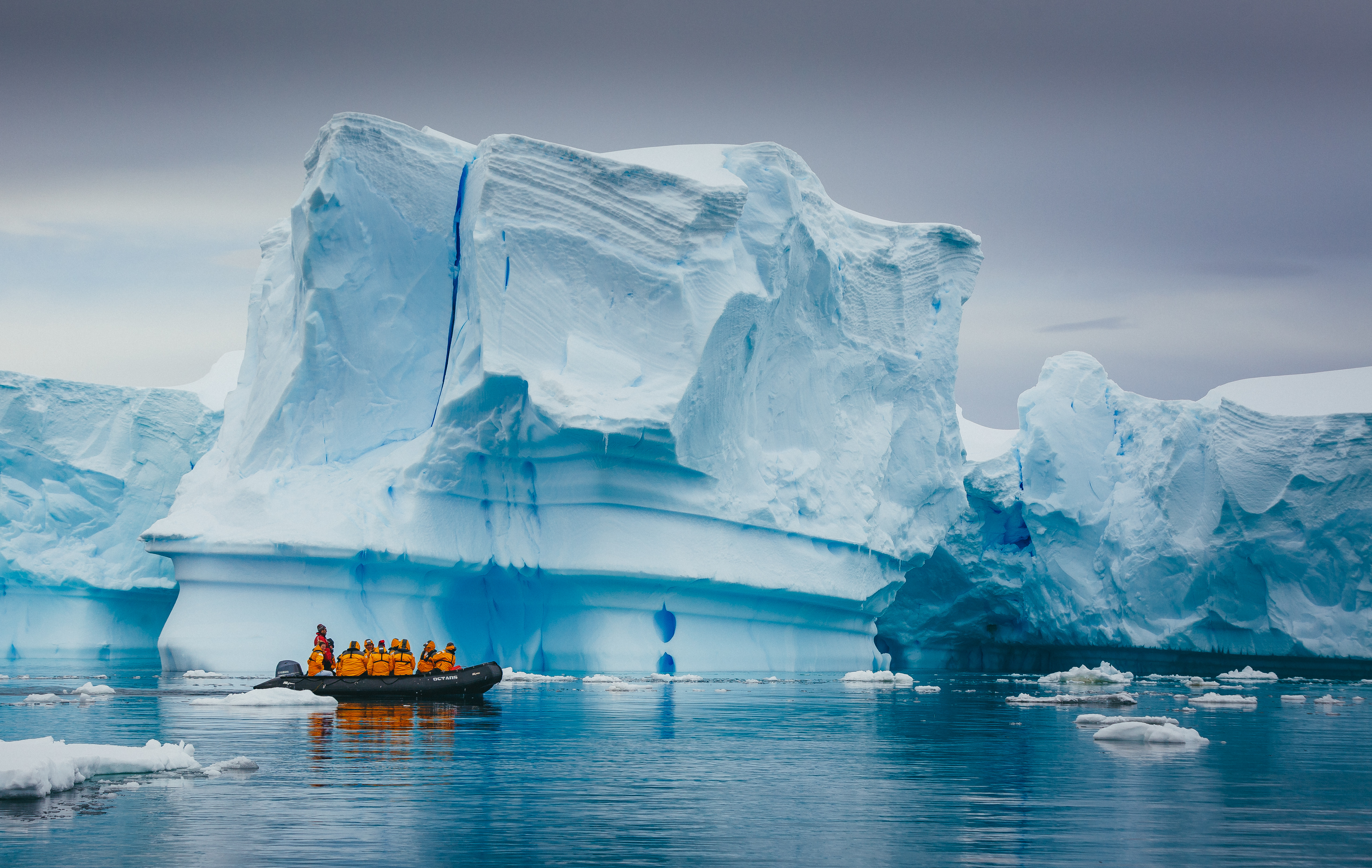 A small Zodiac carries bundled travelers past a towering blue iceberg in the still Antarctic waters below gray skies.