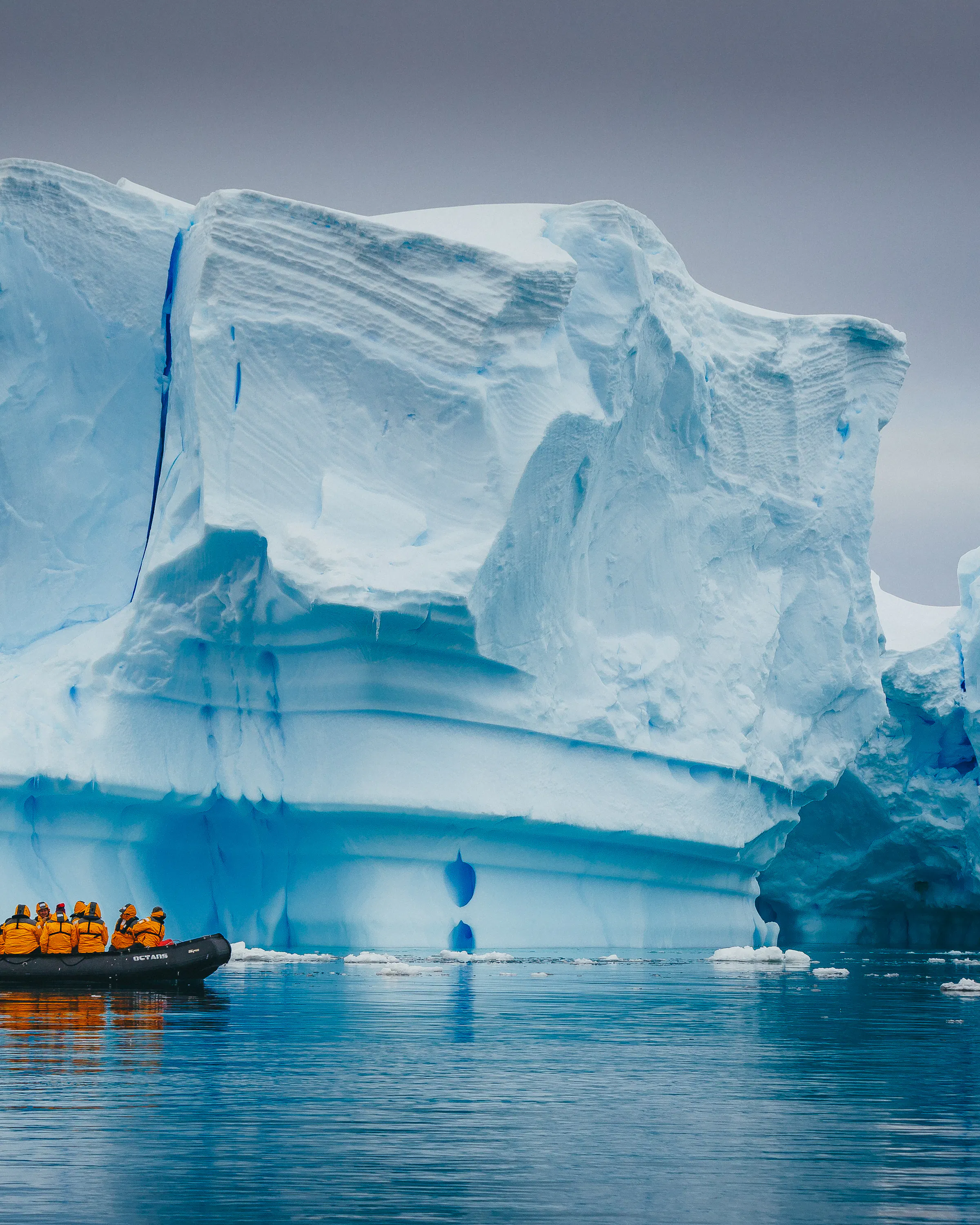 A small Zodiac carries bundled travelers past a towering blue iceberg in the still Antarctic waters below gray skies.