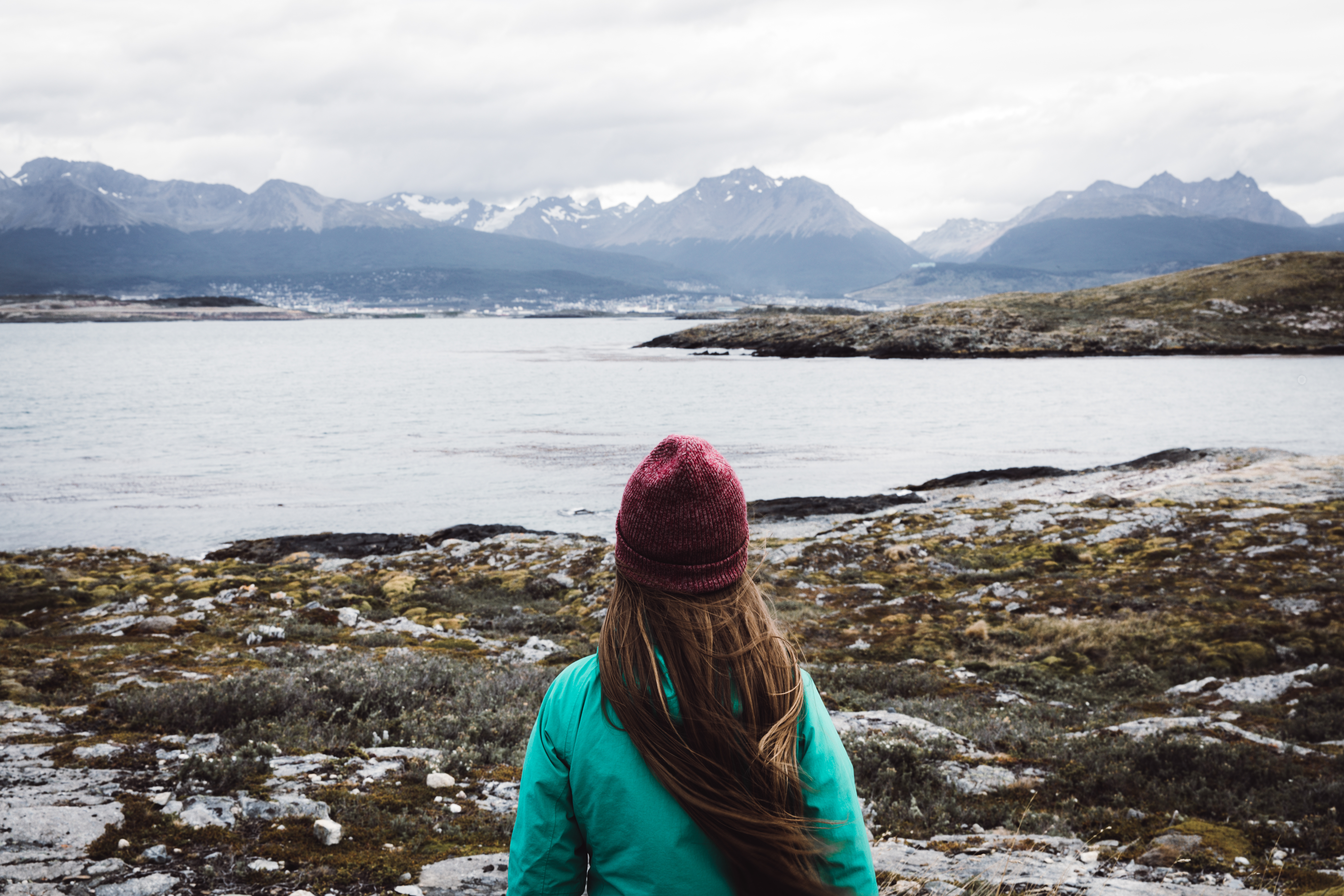 A traveler in a knit cap faces cold waters and rugged peaks, taking in the quiet coastal landscape near Ushuaia.