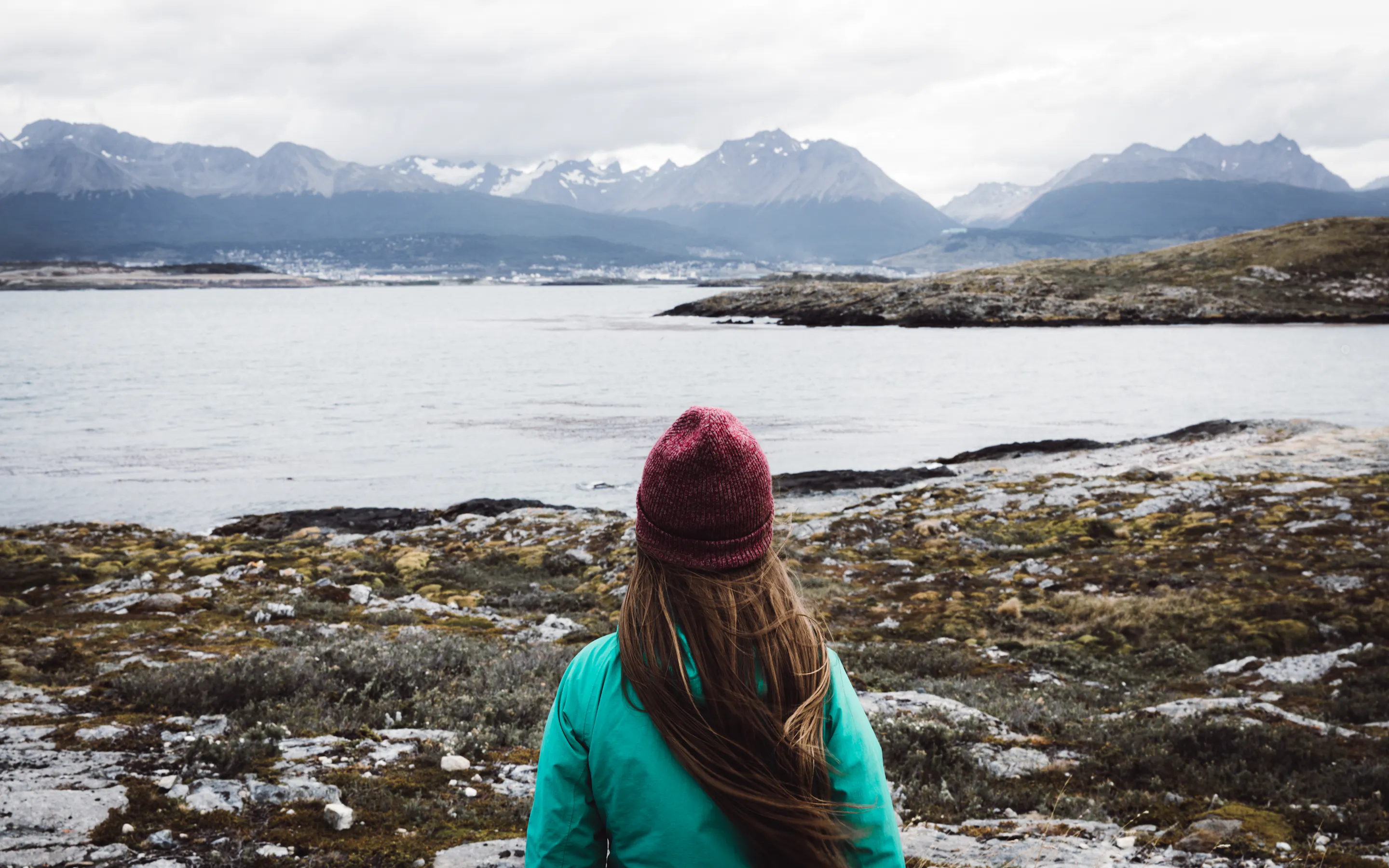 A traveler in a knit cap faces cold waters and rugged peaks, taking in the quiet coastal landscape near Ushuaia.