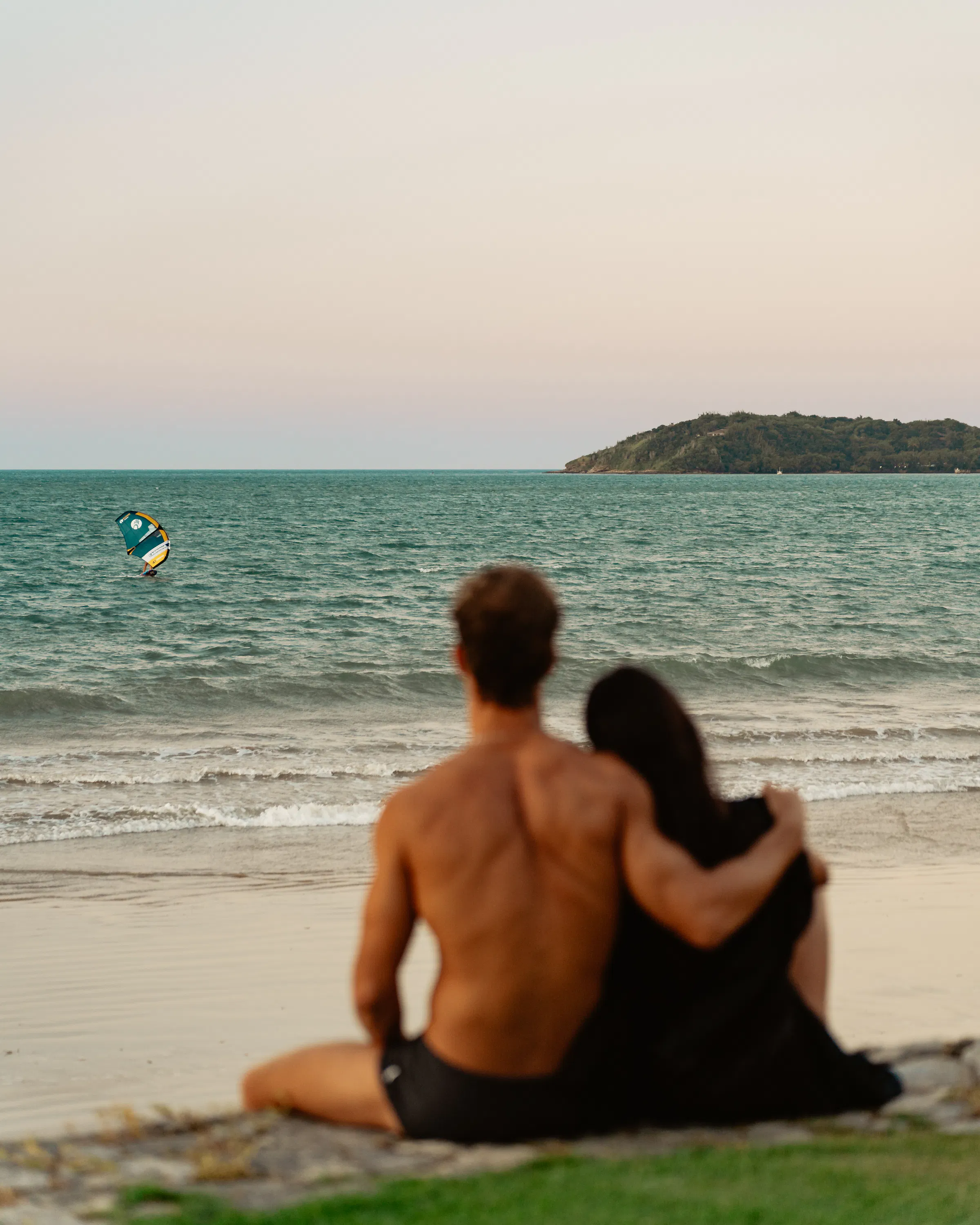A couple sits close on the sand at sunset, watching gentle waves and a kitesurfer off the beach in Buzios.