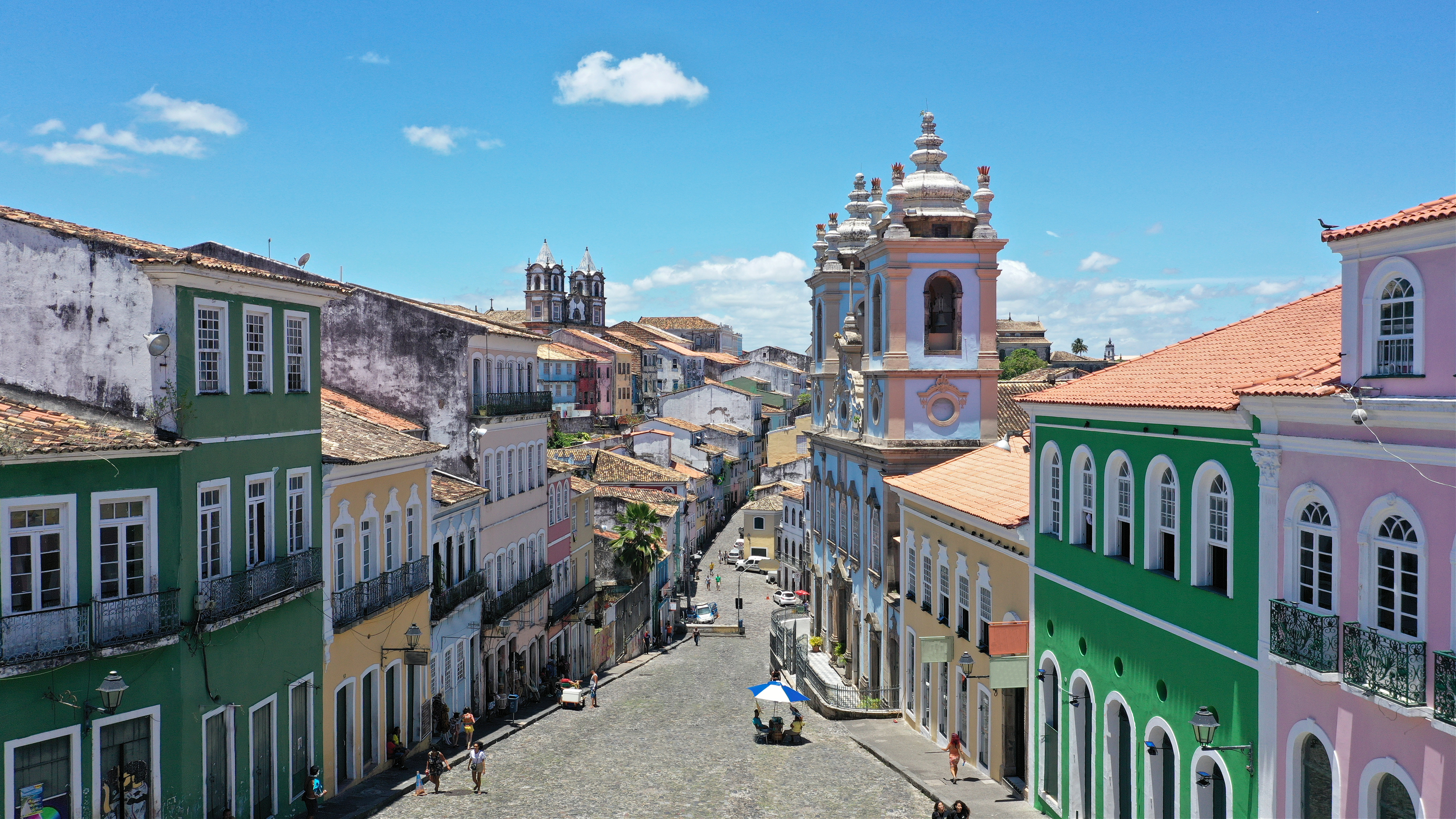A steep colonial street lined with colorful facades climbs through Salvador, with church towers visible beyond.