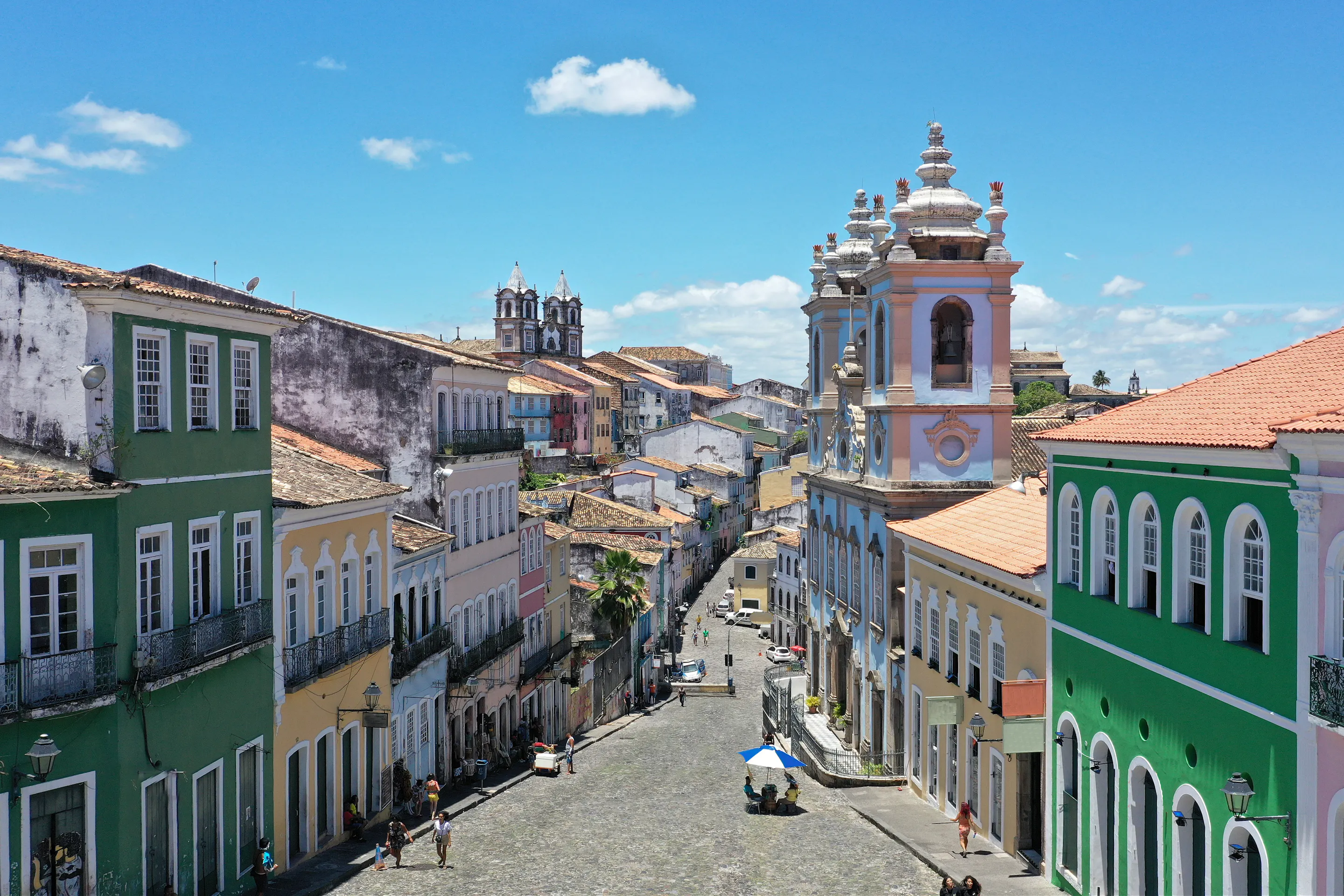 A steep colonial street lined with colorful facades climbs through Salvador, with church towers visible beyond.
