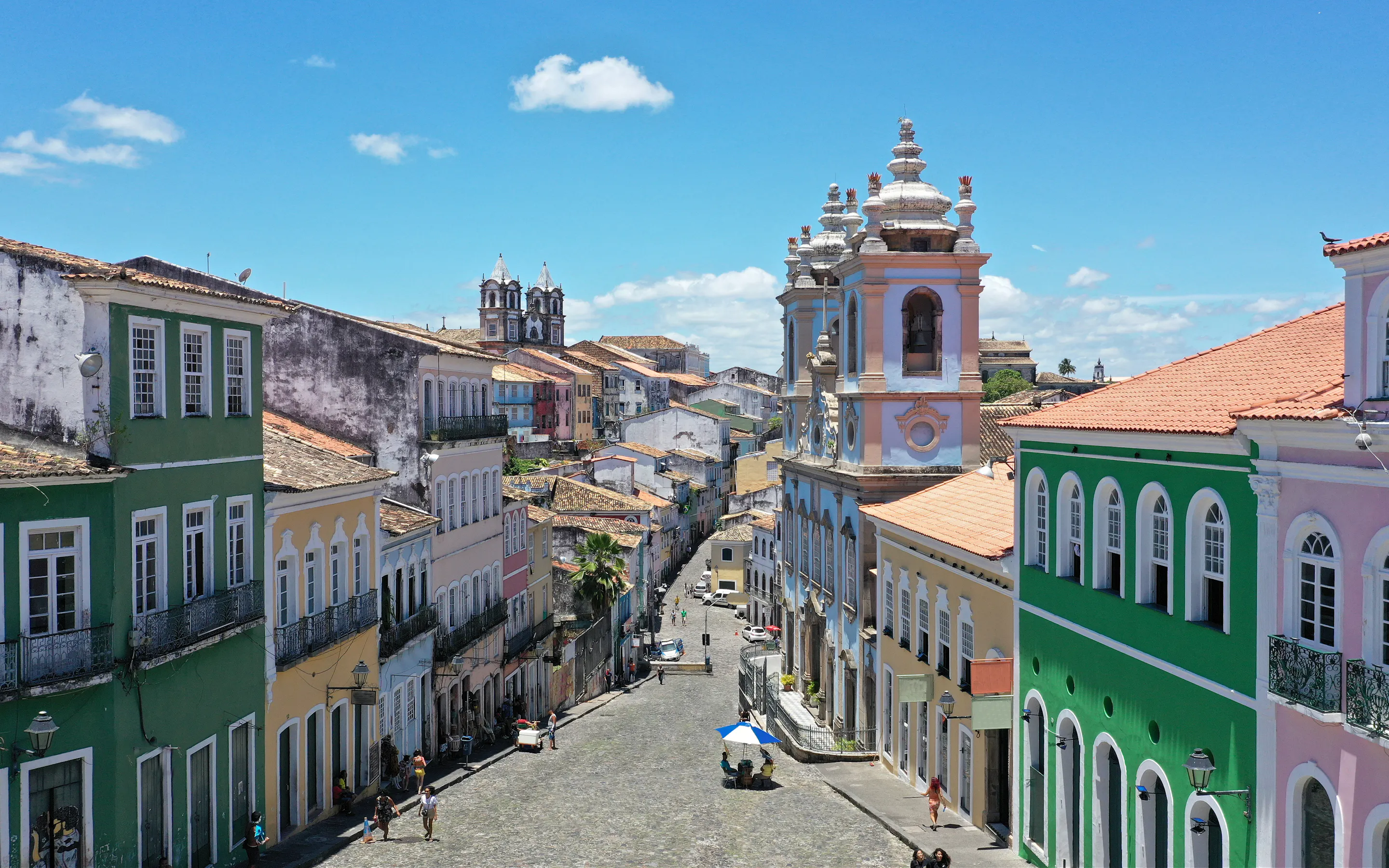 A steep colonial street lined with colorful facades climbs through Salvador, with church towers visible beyond.