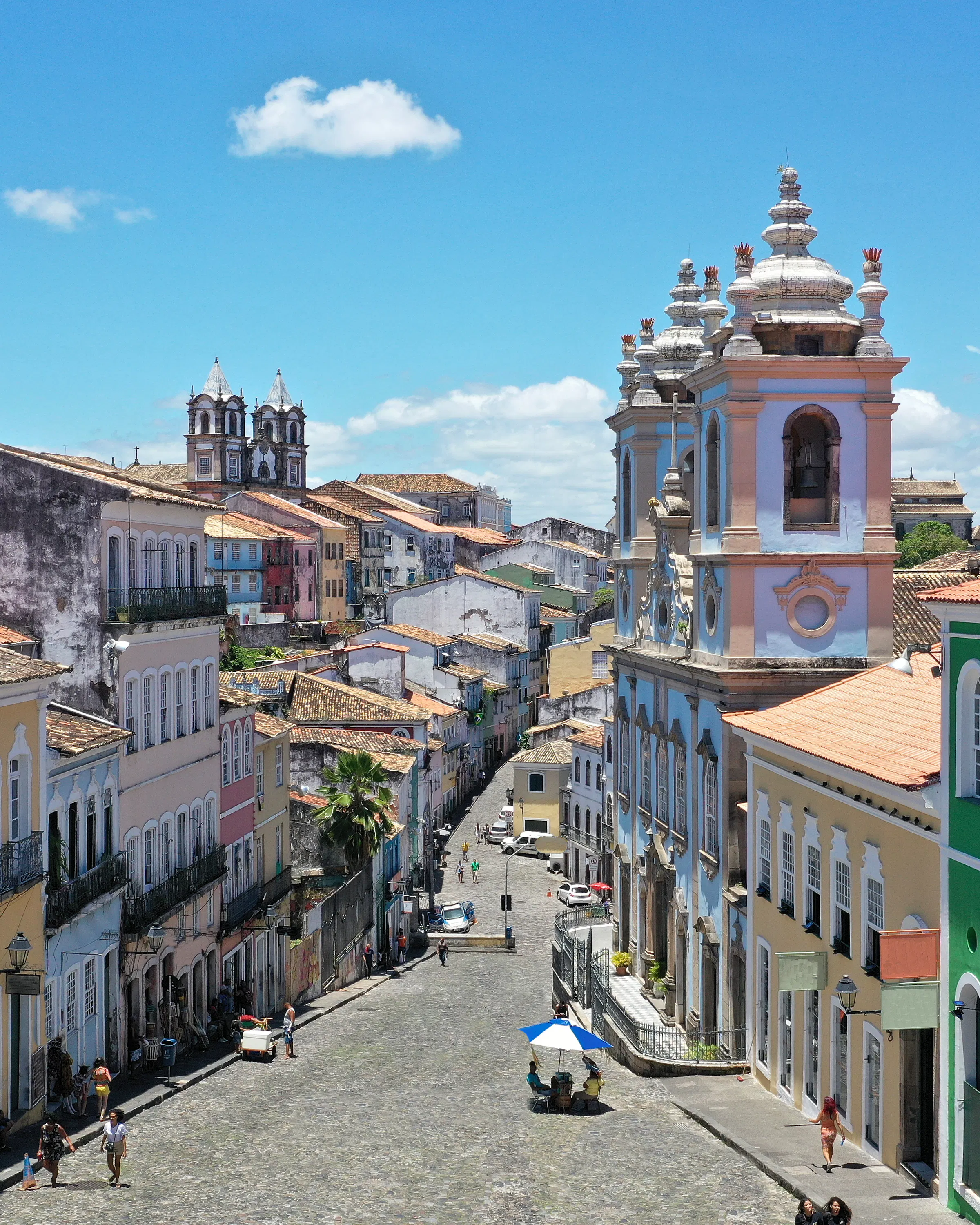 A steep colonial street lined with colorful facades climbs through Salvador, with church towers visible beyond.