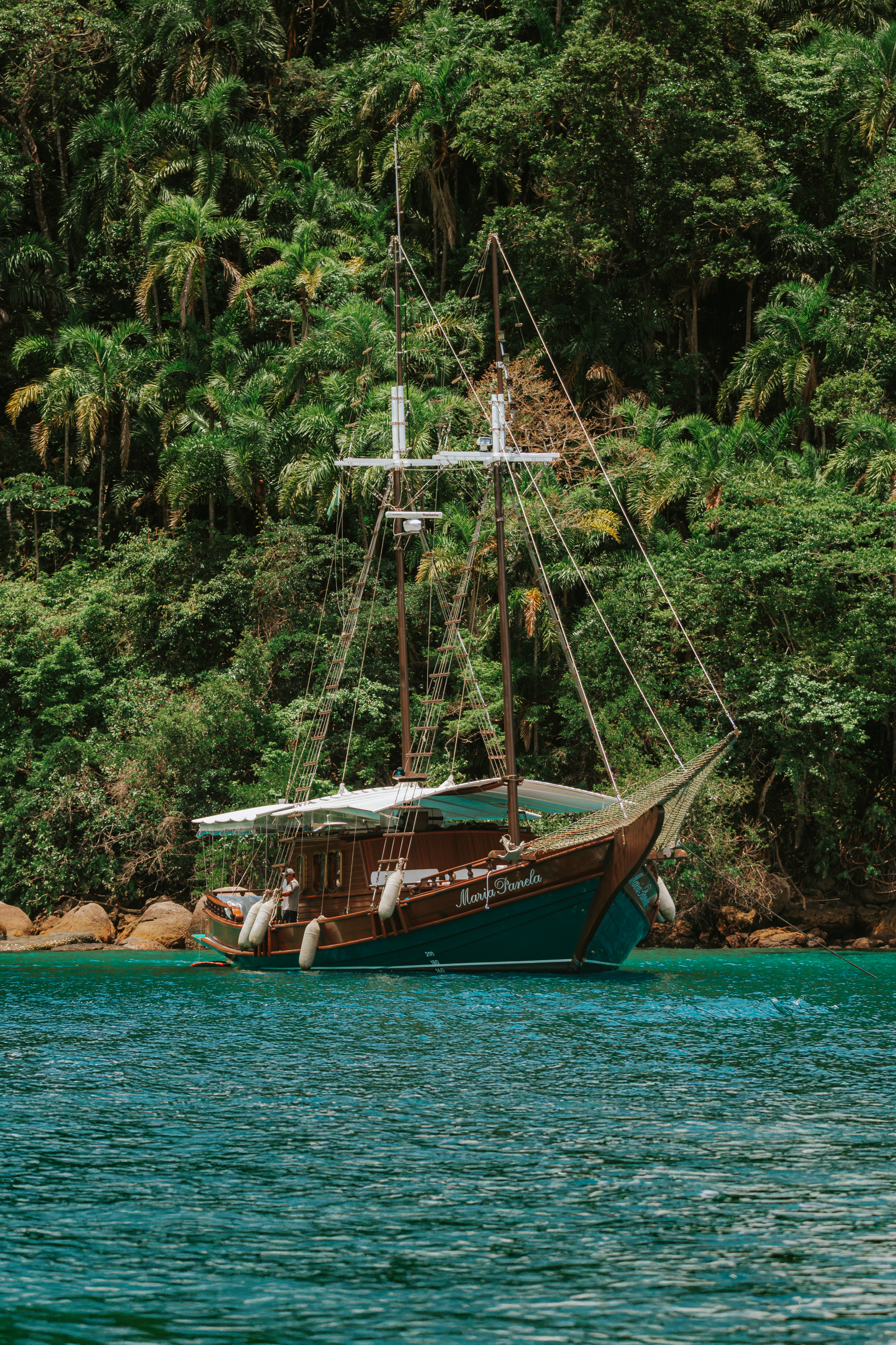A classic schooner drifts on emerald water off Paraty, backed by dense Atlantic forest on the Green Coast.