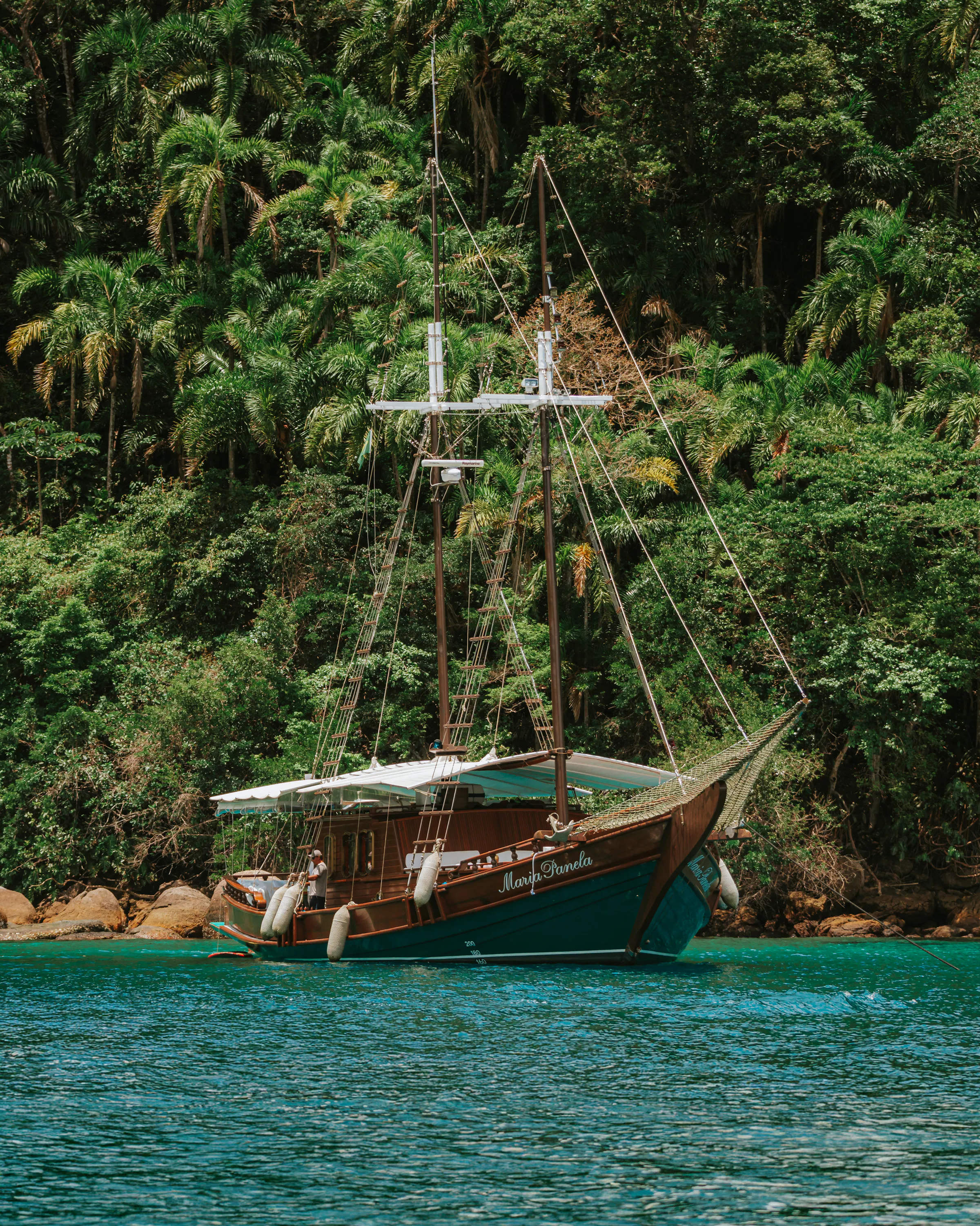 A classic schooner drifts on emerald water off Paraty, backed by dense Atlantic forest on the Green Coast.
