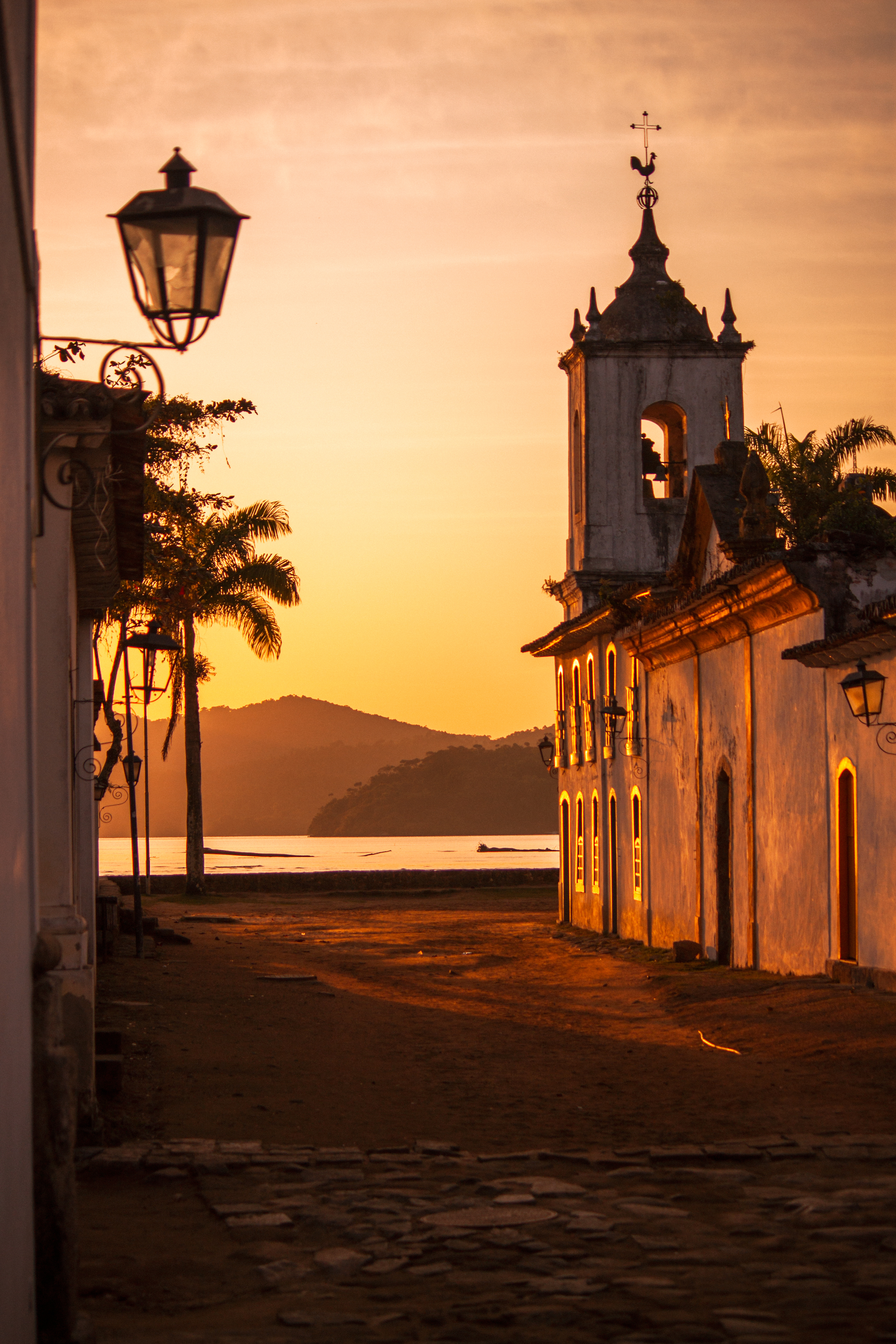 A colonial church glows at sunset in Paraty, with still bay waters and palm silhouettes beyond the empty street.
