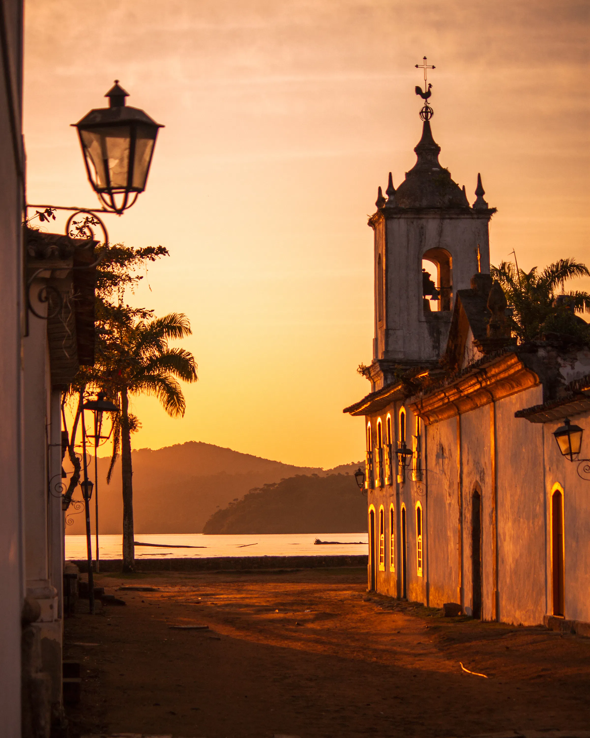 A colonial church glows at sunset in Paraty, with still bay waters and palm silhouettes beyond the empty street.