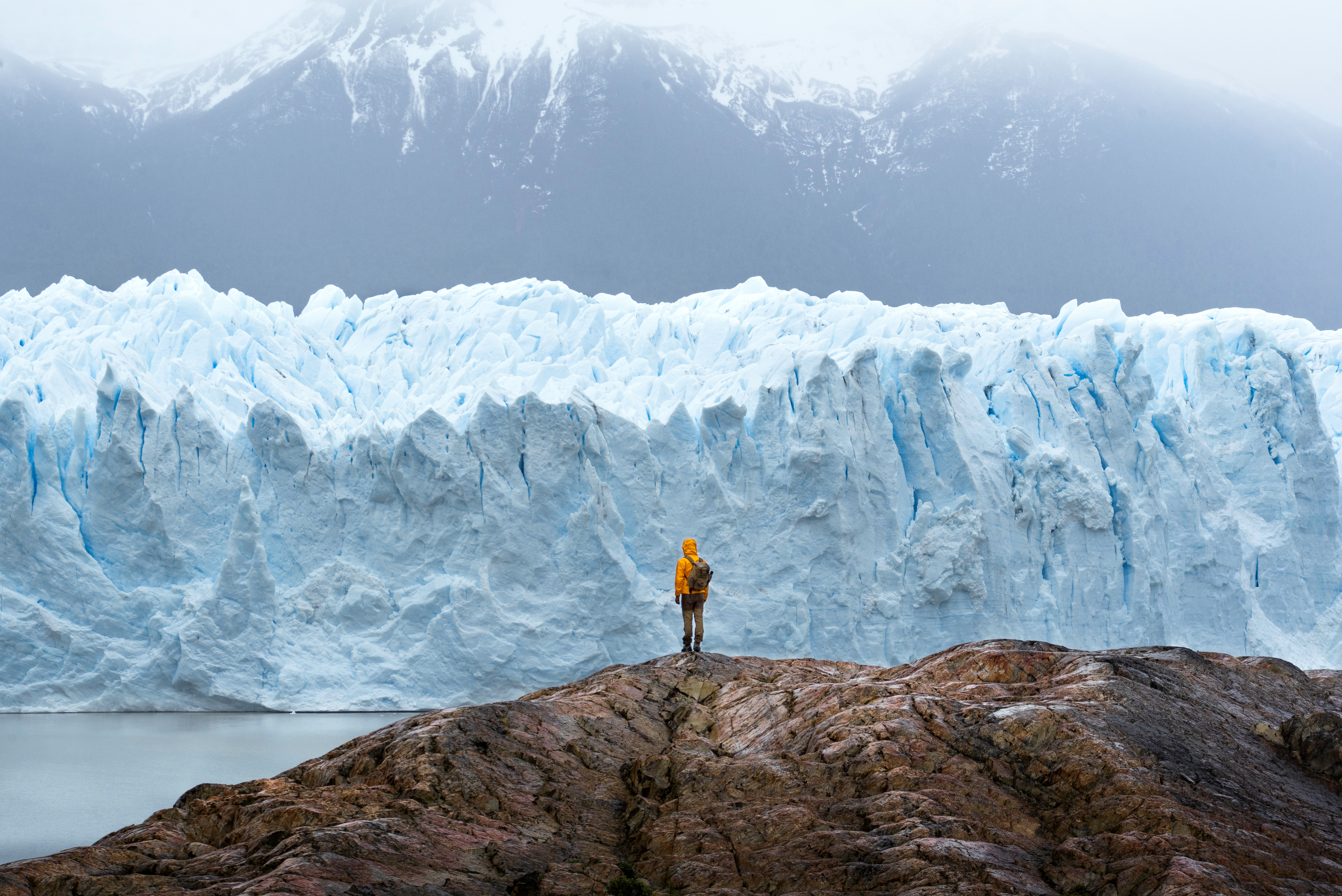 A lone traveler faces a towering blue glacier in Argentine Patagonia, dwarfed by ice walls and dark mountain slopes.