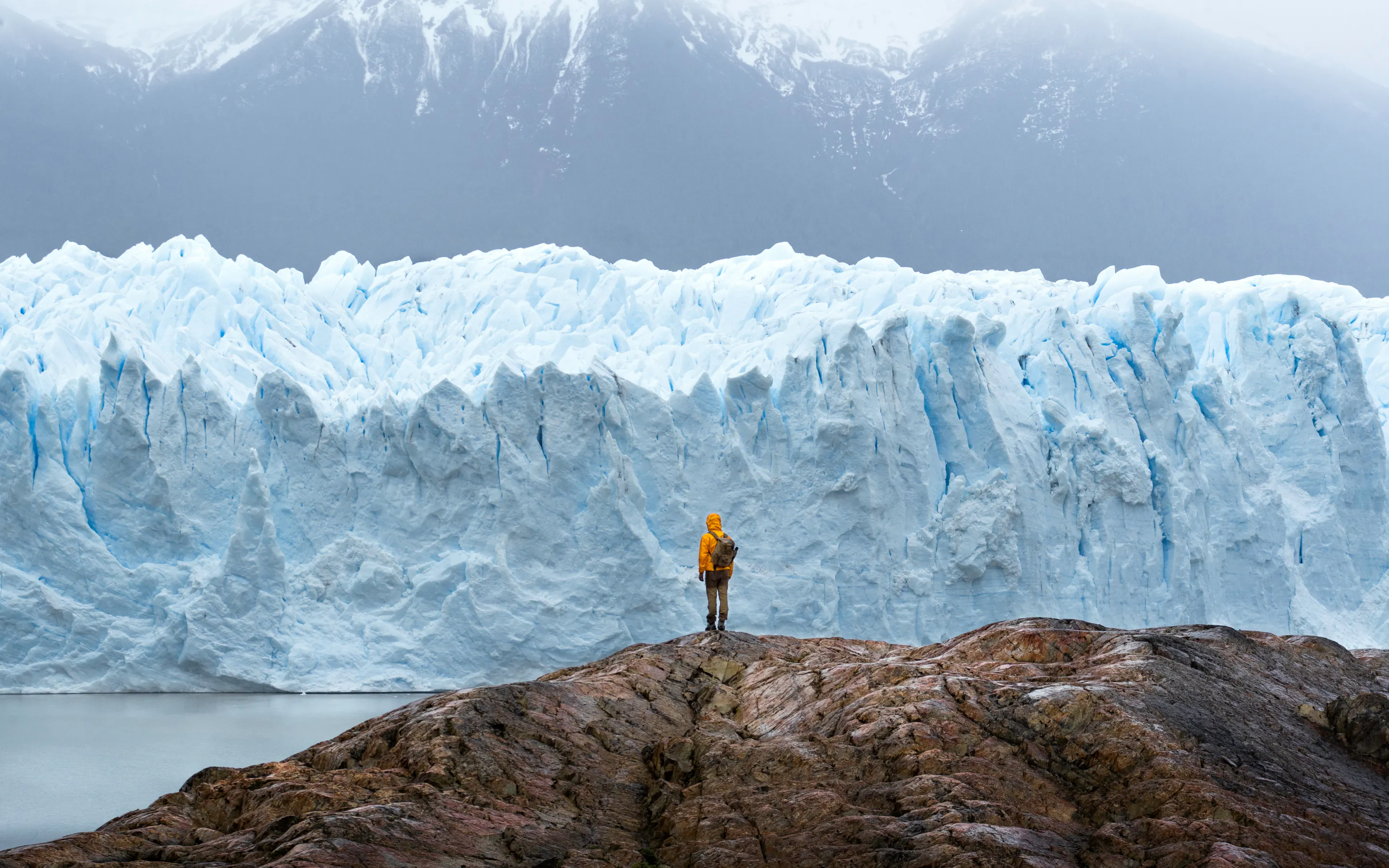 A lone traveler faces a towering blue glacier in Argentine Patagonia, dwarfed by ice walls and dark mountain slopes.