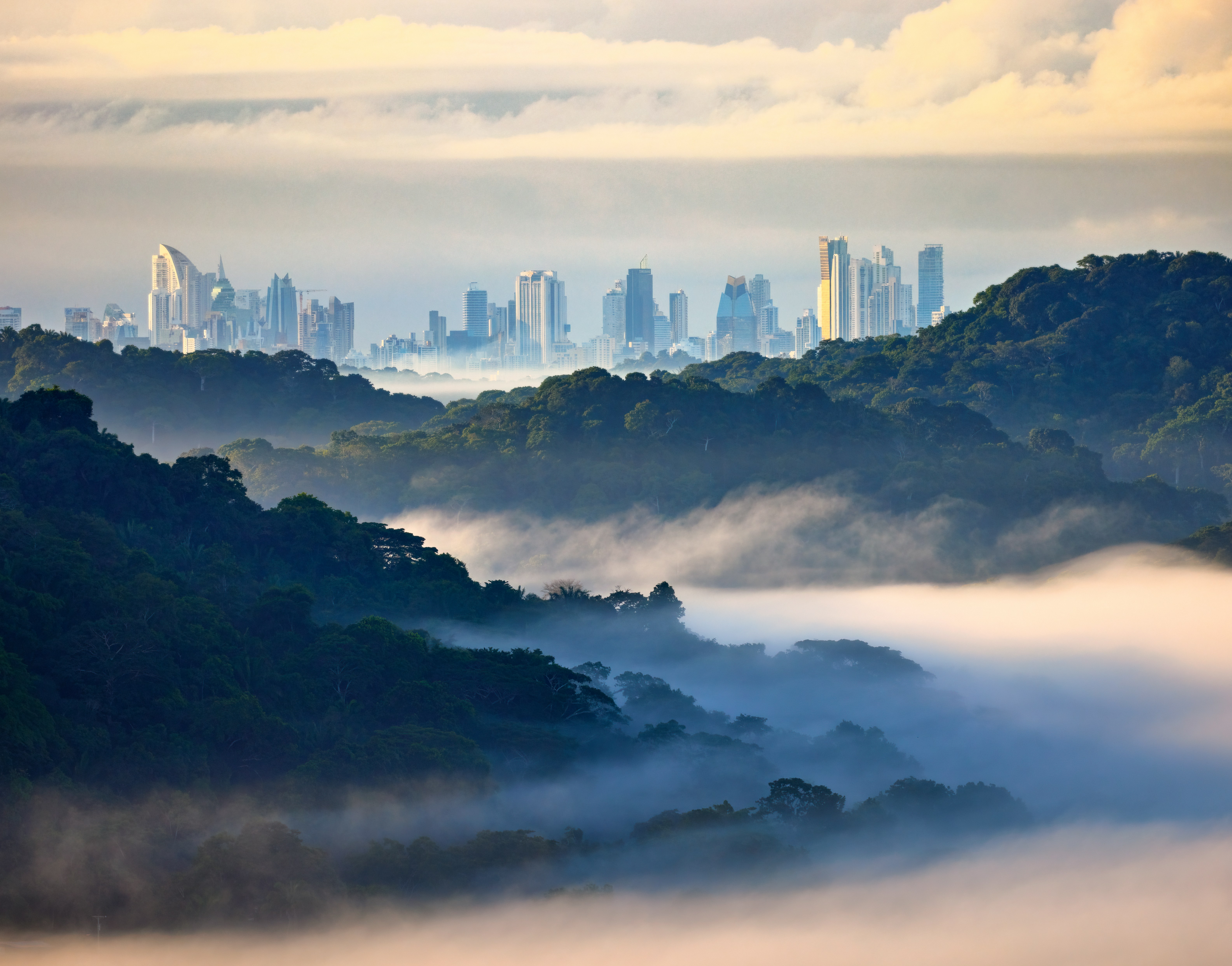 Mist drifts above dense green hills as Panama City's skyline rises in the distance beneath a soft golden sky.
