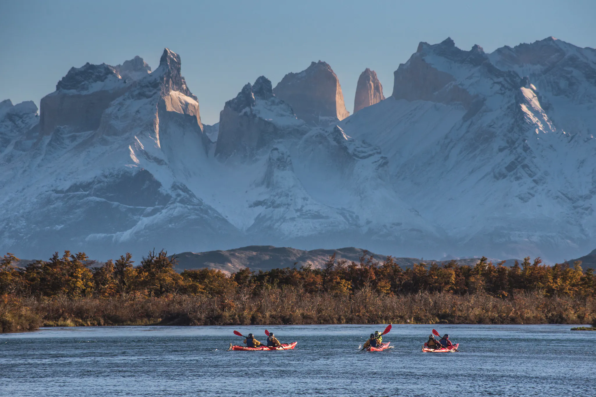Kayakers paddle across a cold blue lake below the jagged Torres del Paine peaks, with autumn trees along the shore.