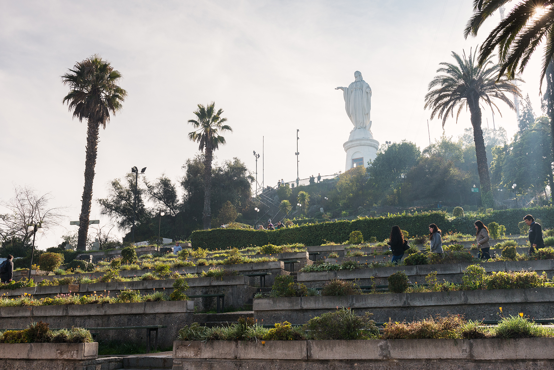 Paths and terraced gardens lead toward the San Cristobal statue in Santiago, framed by palms and soft haze.