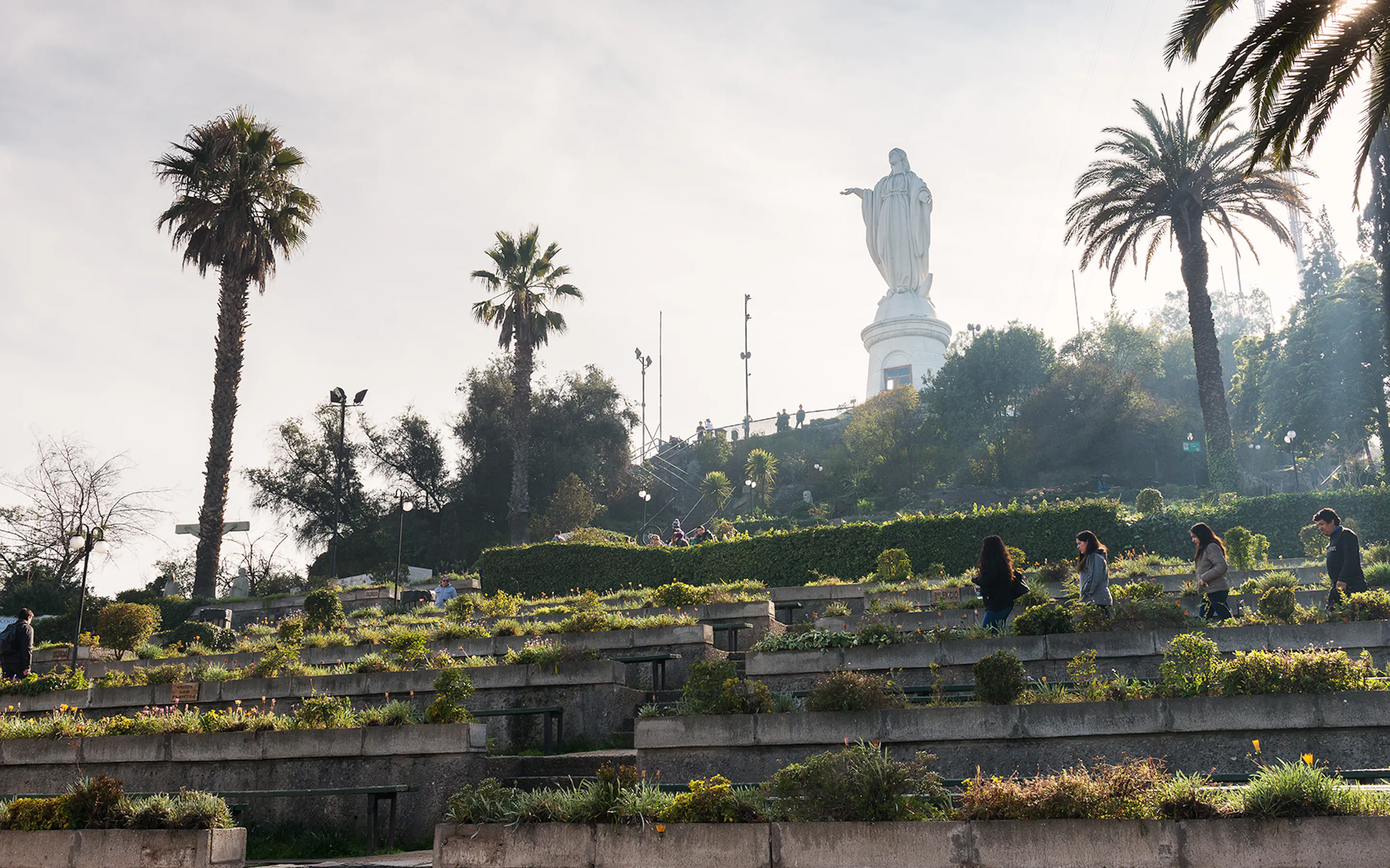 Paths and terraced gardens lead toward the San Cristobal statue in Santiago, framed by palms and soft haze.