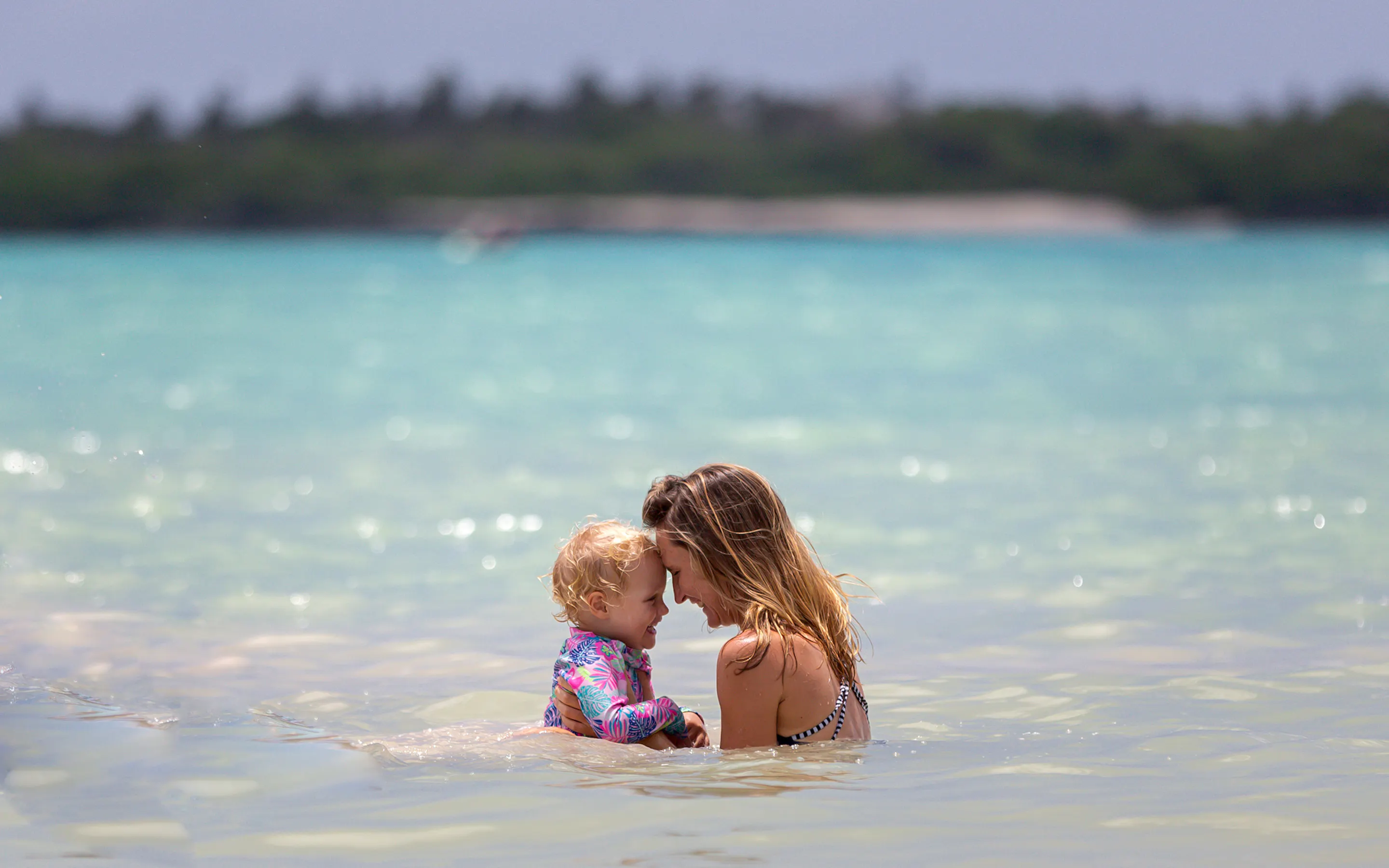 A mother and child smile at each other in shallow turquoise water off the Galapagos coast near Montemar at midday.