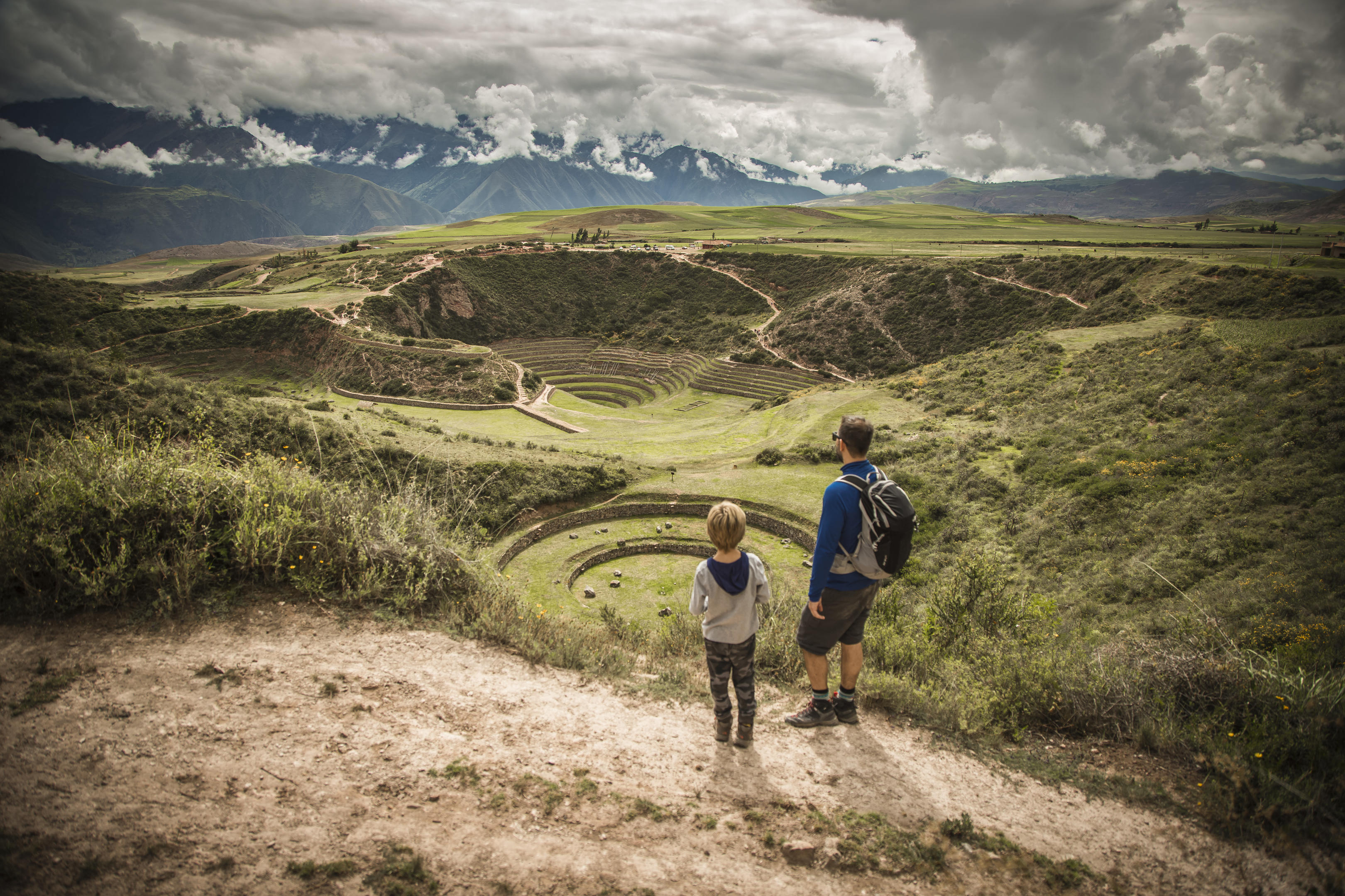 Two guests walk a winding path through the Sacred Valley, with dramatic Andean peaks rising under storm clouds.
