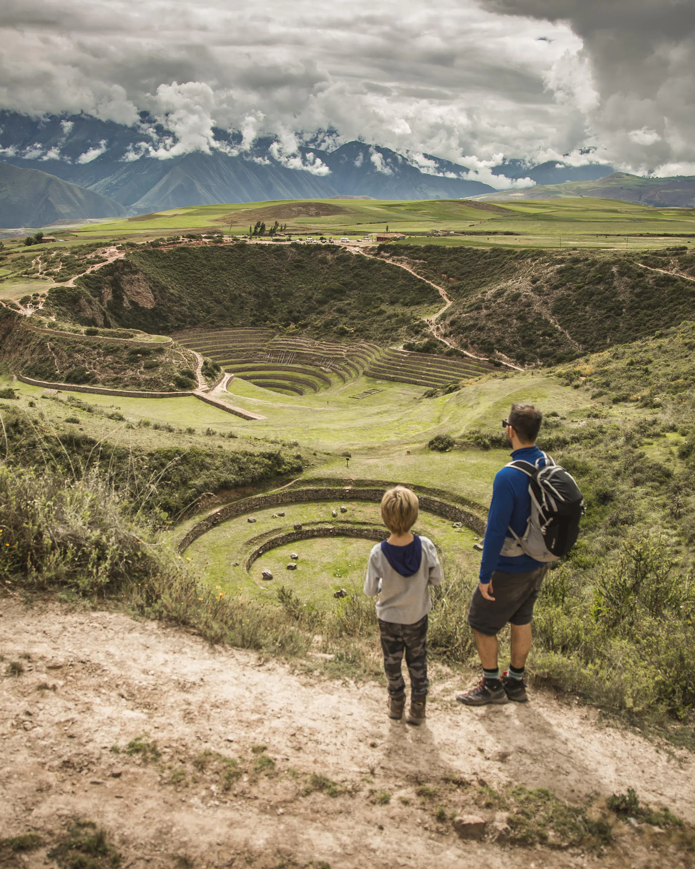 Two guests walk a winding path through the Sacred Valley, with dramatic Andean peaks rising under storm clouds.