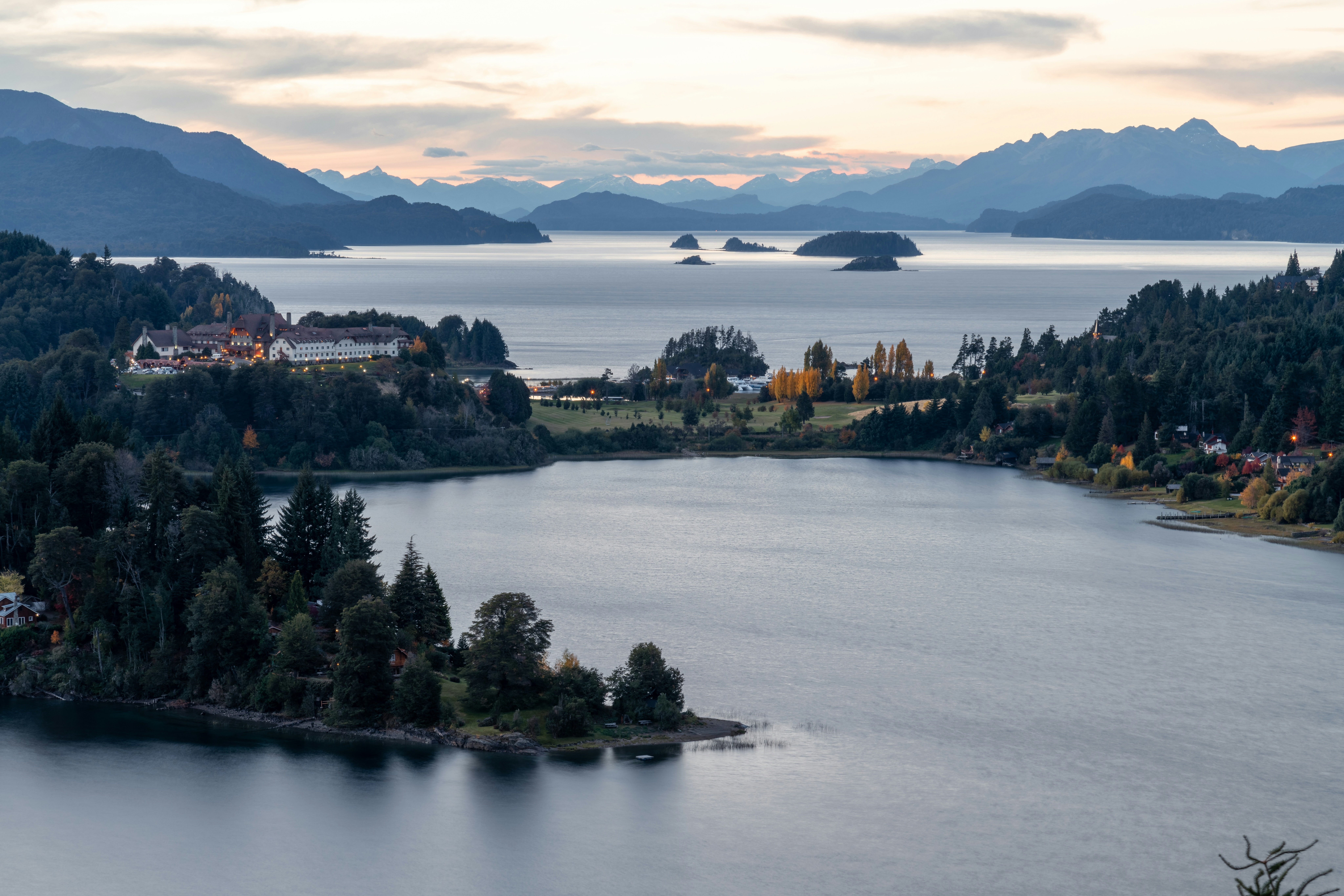 Lakes, wooded peninsulas, and distant islands stretch beneath layered mountains in Bariloche, Argentina, at dusk.