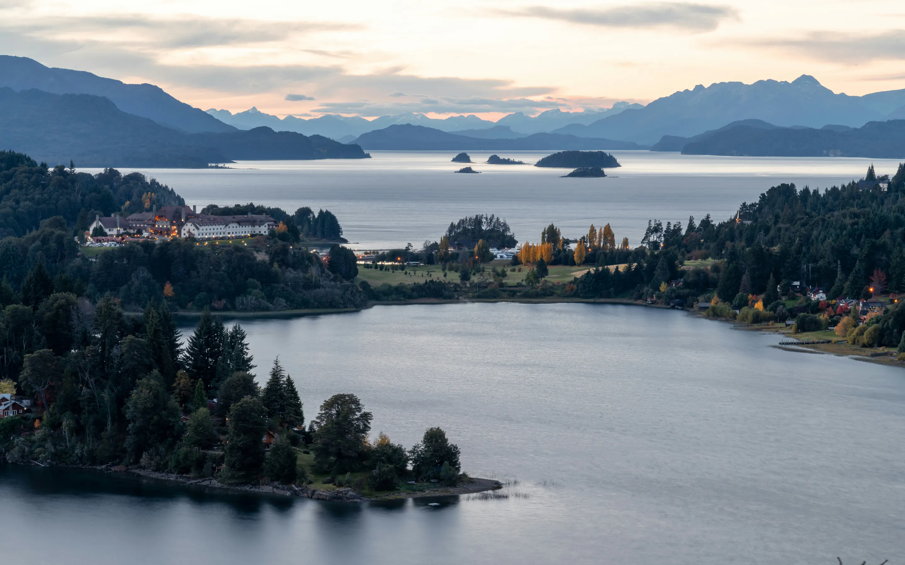 Lakes, wooded peninsulas, and distant islands stretch beneath layered mountains in Bariloche, Argentina, at dusk.
