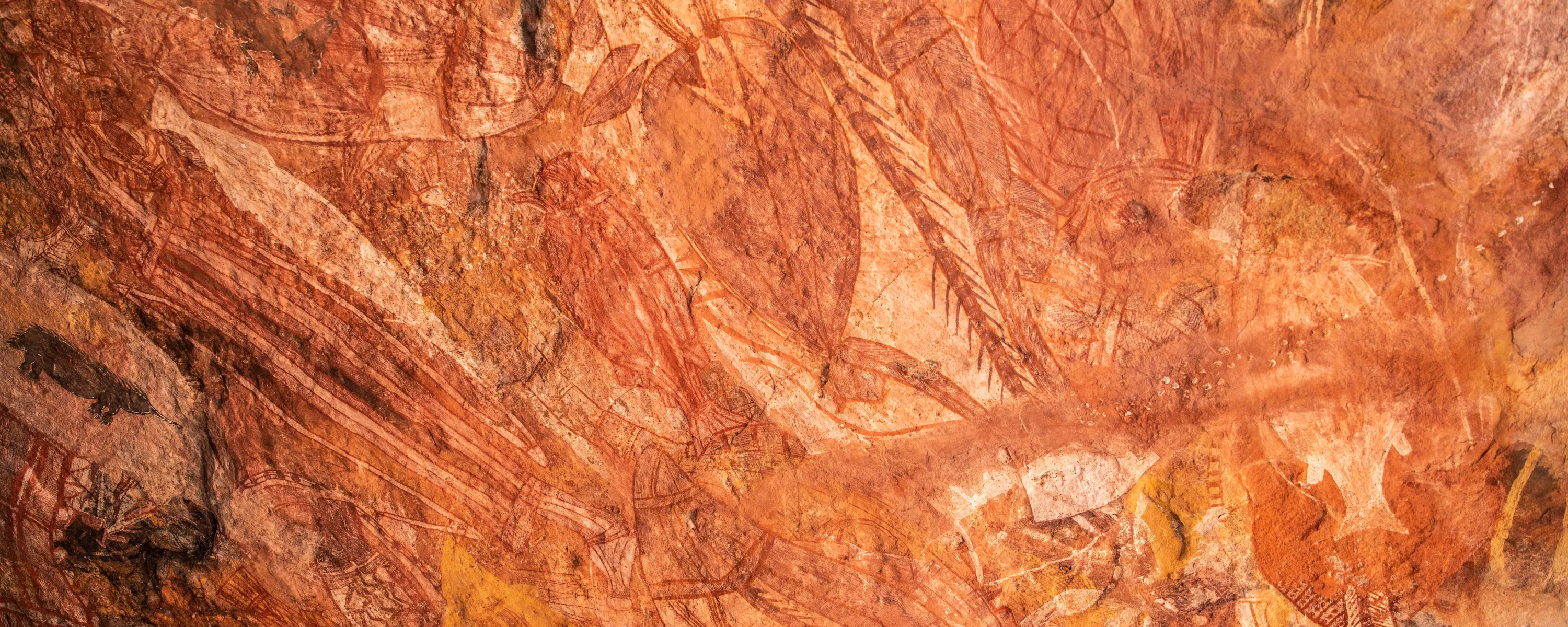 Ancient Aboriginal rock art painted in earthy red and orange tones, depicting animals and figures on a textured stone wall.