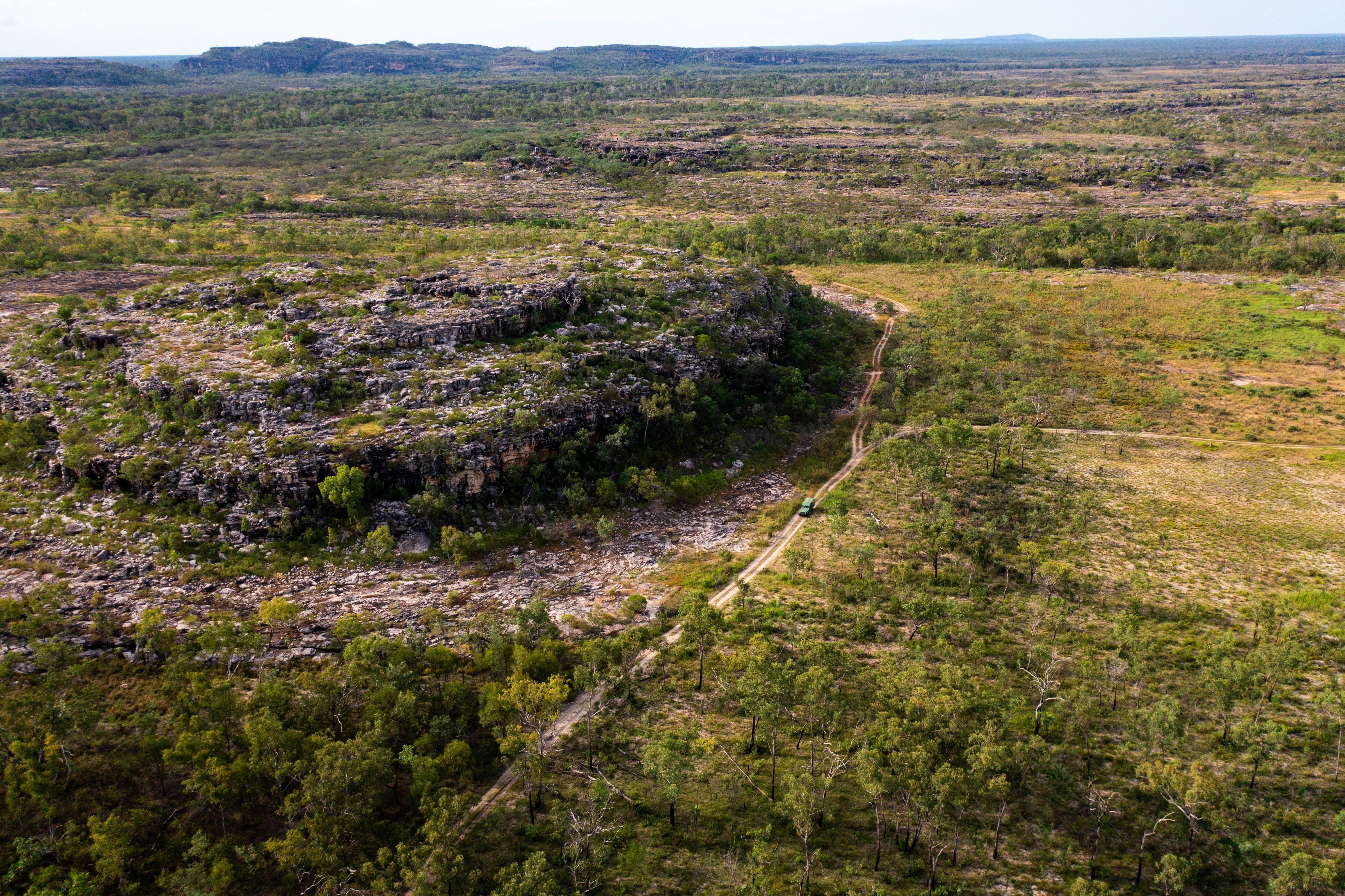 Wide aerial view of a rugged, rocky escarpment surrounded by open woodland and bushland, with a dirt track cutting through the terrain.