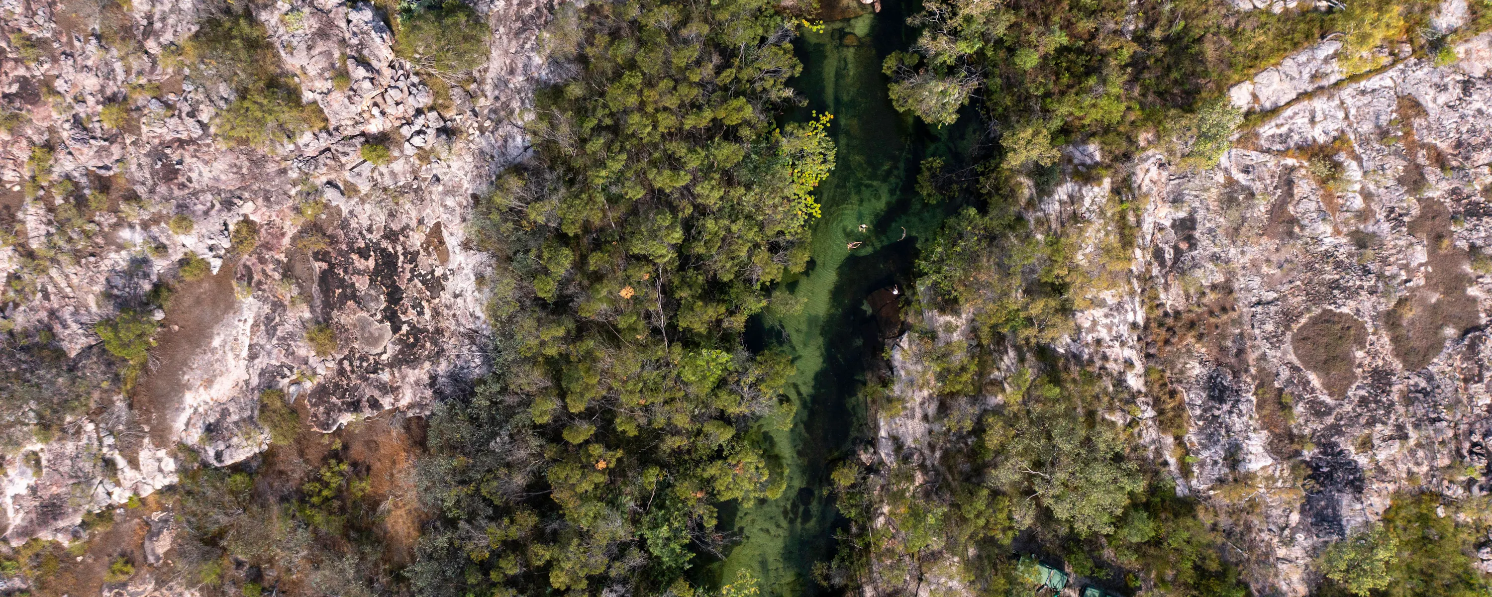 Aerial view of a narrow, green waterhole winding through rocky terrain and trees, with small structures visible nearby.